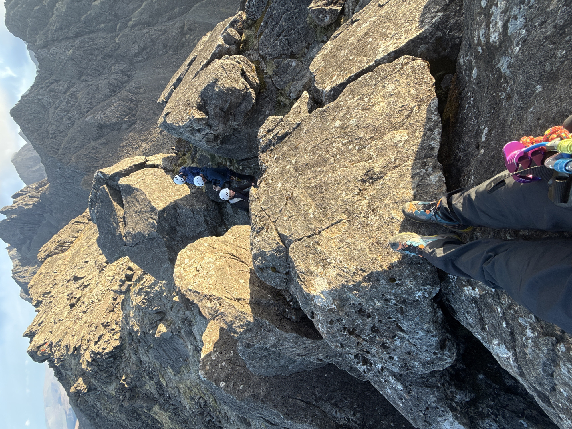 The image depicts a rocky mountain scene. Three people wearing helmets are situated between large rock formations. A person in the foreground is standing on a rock, with their feet visible and climbing equipment attached to their harness. The mountain cliff is visible in the background. The location is Unknown, Unknown.