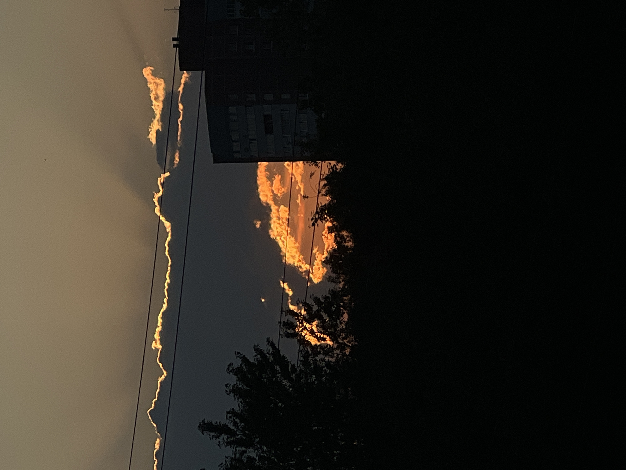 The image captures a sunset over Saint Petersburg, Russia, characterized by dramatic lighting and atmospheric conditions. The sky is a gradient of warm oranges and yellows, with the sun's rays piercing through dark clouds, creating a halo effect. A building, likely residential, is silhouetted against the sky on the right side of the frame. In the foreground, the dark silhouettes of trees and power lines suggest an urban or suburban setting. The overall impression is one of a serene yet powerful natural phenomenon observed within a cityscape.