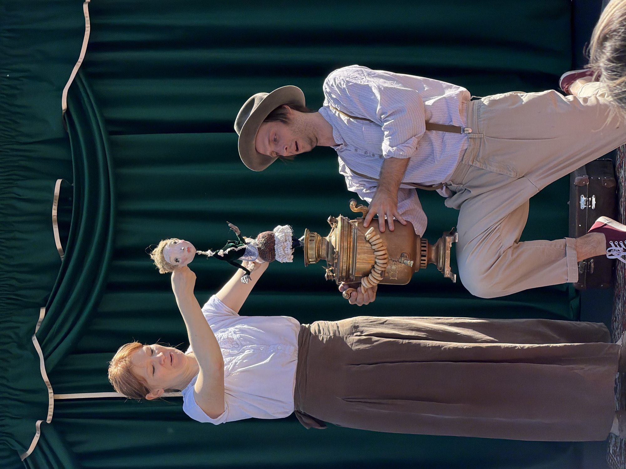 The image captures an outdoor theatrical performance against a dark green curtain backdrop, a scene reportedly taking place in Khimki, Russia. A woman, estimated to be in her 30s or 40s, with reddish-brown hair, stands on the left. She wears a white short-sleeved blouse with lace detailing and a long, pleated light brown skirt tied at the waist. Her right arm is extended, holding the head of a blonde, pale-skinned puppet over a large, ornate brass samovar. The puppet's body, dressed in a dark green top and a frilly brown and white skirt, dangles below the head. On the right, a man, also appearing to be in his 30s, kneels on his right knee. He wears a light-colored fedora, a light blue and white striped long-sleeved shirt with brown suspenders, and light beige cuffed trousers with red sneakers. He holds the brass samovar with both hands, looking upward and to the left with a slightly open mouth, indicating engagement or surprise. A string of light-colored, ring-shaped pastries is draped around the samovar. The ground is partially covered by a patterned rug on which a small, brown vintage-style suitcase with metal clasps rests behind the kneeling man. The scene is brightly lit by natural sunlight, casting visible shadows. The top of a person's head is visible in the bottom right corner, suggesting an audience.