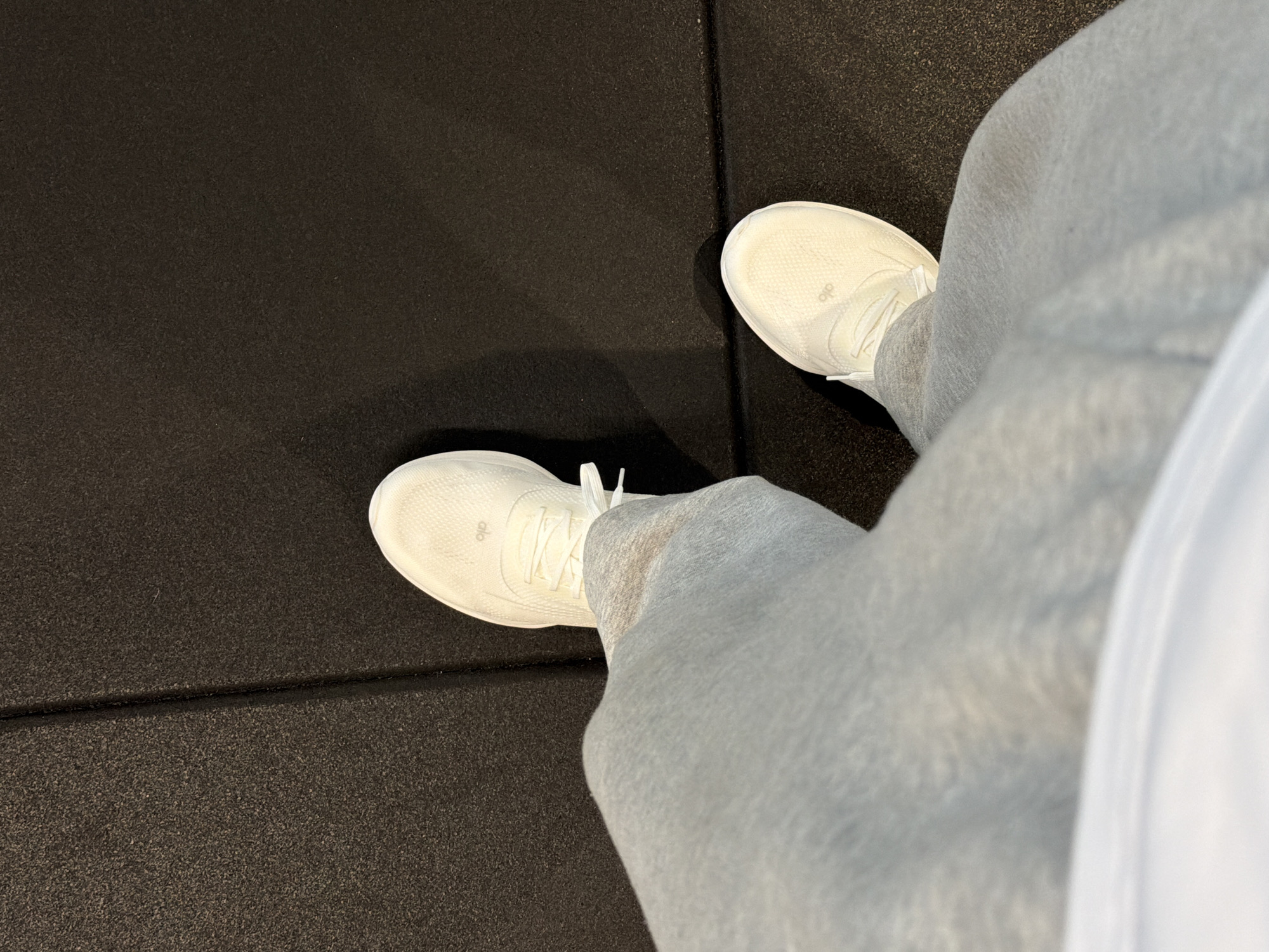 The image shows a person's perspective looking down at their feet, which are clad in white athletic shoes from the brand "Alo" and grey sweatpants. The person is standing on a black, textured surface with linear indentations, suggesting a rubber or gym floor. The setting is likely an indoor location, such as a gym or fitness studio. There is no indication as to where in Los Angeles this photo was taken.