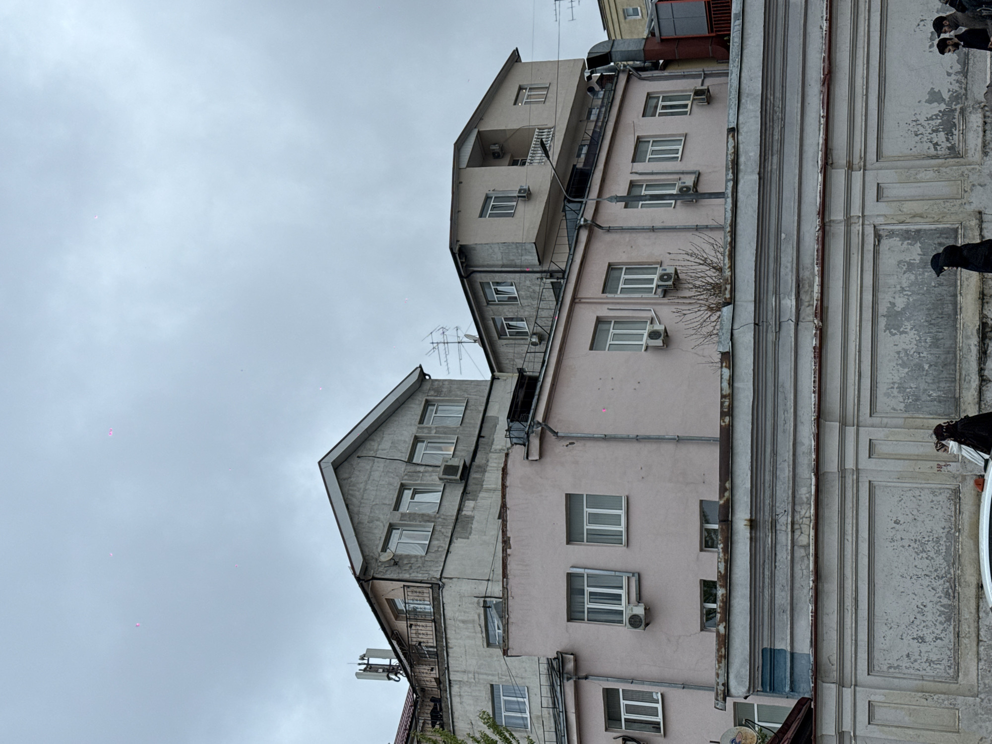 This media file captures an urban street scene in Moscow, Russia, featuring a dense arrangement of multi-story buildings under an overcast sky. The image is rotated, presenting the buildings vertically.
In the foreground, on what would be the right side of an upright image, there is an older building with a worn, textured facade displaying decorative vertical panels and peeling paint. A white car is partially visible at the bottom, and several individuals wearing dark clothing and hats or hoods are partially seen standing near the car and the building. A faint red number "38" (or "38p") is visible on the wall near them.
Behind this older structure, a light pink or peach-colored building rises, characterized by numerous rectangular windows and visible air conditioning units attached to its exterior. Further back and to the left, another building with a grey, possibly concrete or stuccoed finish, features multiple windows, balconies, and air conditioning units, topped with a distinctive sloped roofline. Beyond these, a taller, lighter beige building is also visible.
The sky is uniformly overcast and light grey, with several small, bright pink dots scattered across it, which appear to be lens flares or digital artifacts rather than actual objects. Bare tree branches are visible near the pink building, suggesting a colder season. Various pipes and overhead wires are discernible between and around the buildings. The scene depicts a typical daytime urban environment, likely in a colder period given the bare branches and clothing.
