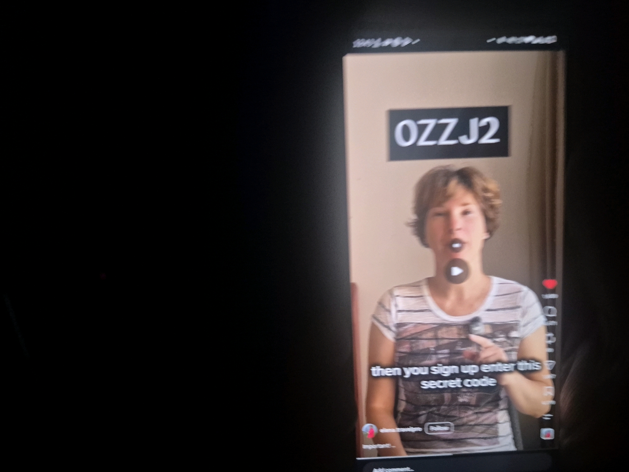 The image shows a close-up of a smartphone screen displaying a video. A woman with short, light brown hair is visible in the video, wearing a striped t-shirt. She is pointing to text on the screen that reads "OZZJ2" and "then you sign up enter this secret code". There is a play button overlaid on her face. The background of the video is a plain, light-colored wall. The smartphone itself is held by a hand, parts of which are visible on the right side of the image. The overall environment appears to be indoors, given the lighting and the presence of a device screen. There is no identifiable information in the image that places it in Al Muzahimiyah, Saudi Arabia.