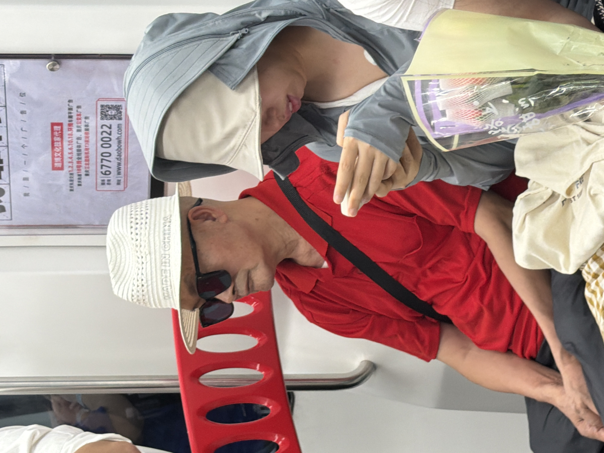 The image depicts two individuals, an elderly man and a younger person, seated in what appears to be a subway car in Yuzhong District, China. The man, on the left, is wearing a red polo shirt, dark sunglasses, and a white woven hat with "MADE IN CHINA" visible. He is looking to his right. The younger person, on the right, is wearing a gray hooded jacket and a light-colored hat, obscuring their face. They are looking down at their hands, which are holding a smartphone and a bouquet of flowers. The subway car interior is visible, with a red railing in the foreground and a white wall with an advertisement behind the individuals. The advertisement features Chinese text and a QR code. The overall lighting suggests an indoor environment.