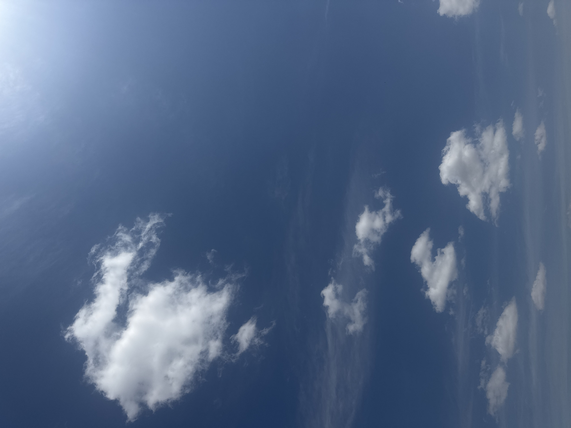The image shows a clear blue sky with scattered white clouds. The sun is visible in the upper right corner, casting a bright light across the scene. There are no people, buildings, or other objects visible in the image, indicating an open sky view, possibly from an outdoor location. The environment appears to be natural and atmospheric, with no specific urban features from село Умбеталы Карибаева, Kazakhstan being depicted.