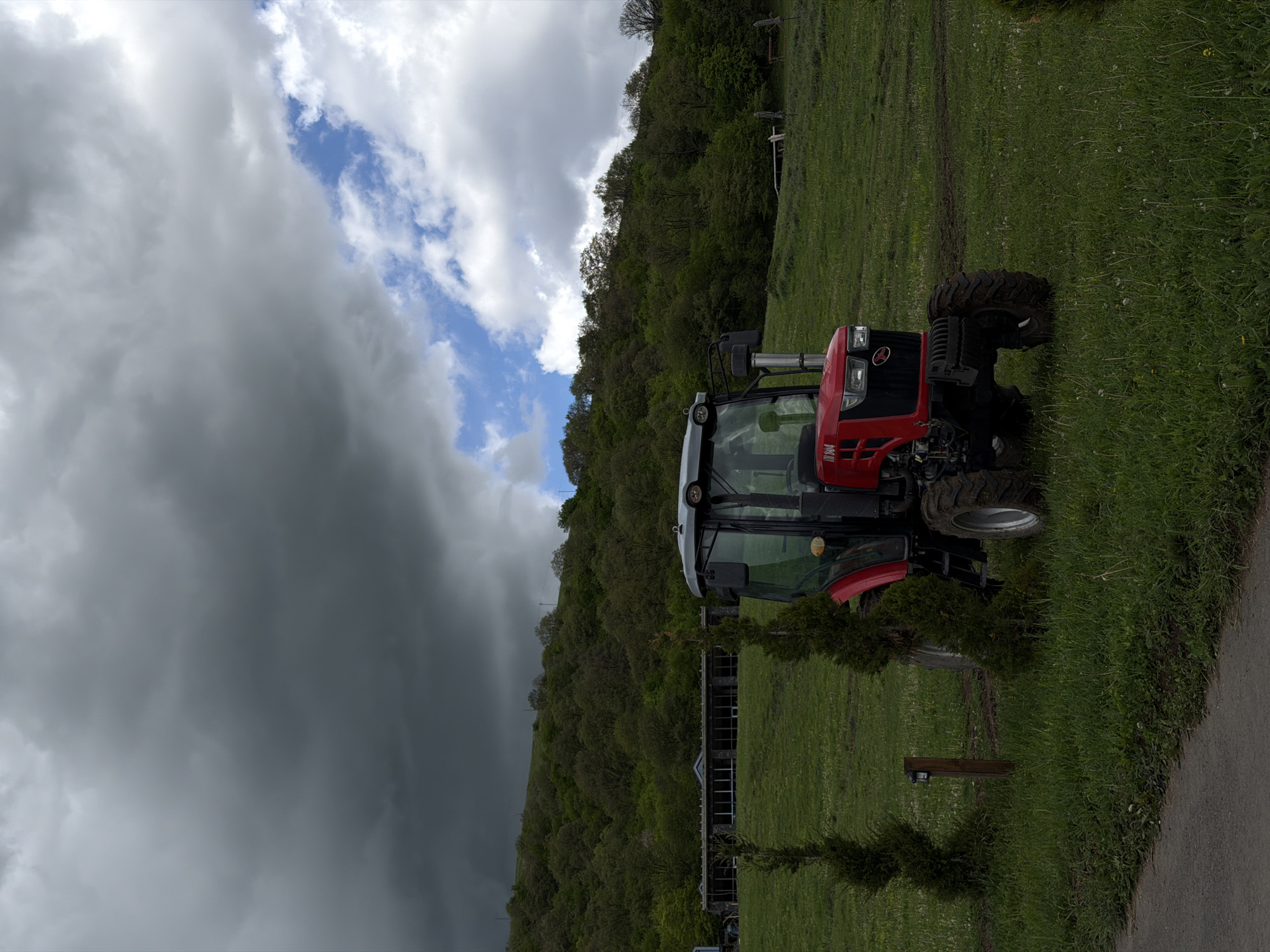 A medium-shot, outdoor photograph depicts a red and gray tractor parked on a grassy field. The field appears to be situated at the base of a densely wooded hillside. Behind the tractor, a long, low building with multiple windows is visible. A small, coniferous tree is planted near the front of the tractor. Above the hillside, a dramatic sky of dark, storm clouds contrasts with patches of blue sky. The scene is in Dsegh, Armenia.