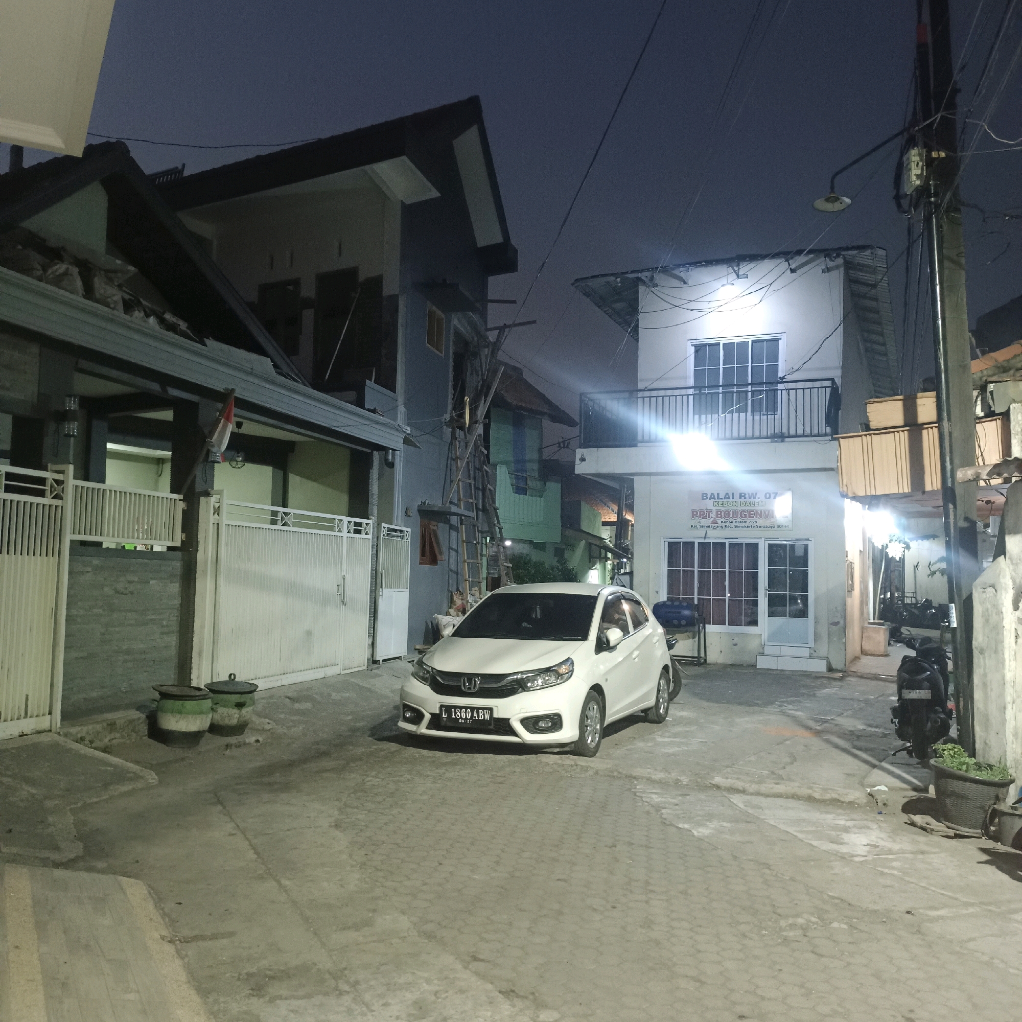 The image depicts a residential street scene at night in the city of Surabaya, Indonesia, specifically at the Balai RW 07, Kebon Dalem, located at Jalan Dalem 7.29, Kel. Simolawang, Kec. Simokerto, Surabaya 60184. A white Honda Brio car with license plate L 1860 ABW is parked in the foreground on a paved street. To the left of the car is a house with a white fence and a partially visible Indonesian flag hanging from the roofline. Adjacent to this house is a multi-story building with a balcony on the upper floor. On the right side of the street, another building displays a sign for "BALAI RW. 07 KEBON DALEM." A dark-colored motorcycle is parked near this building. The environment is lit by streetlights and internal building lights, casting shadows on the ground. Two large trash bins with green and black markings are situated near the white fence on the left. The sky is dark, indicating nighttime.