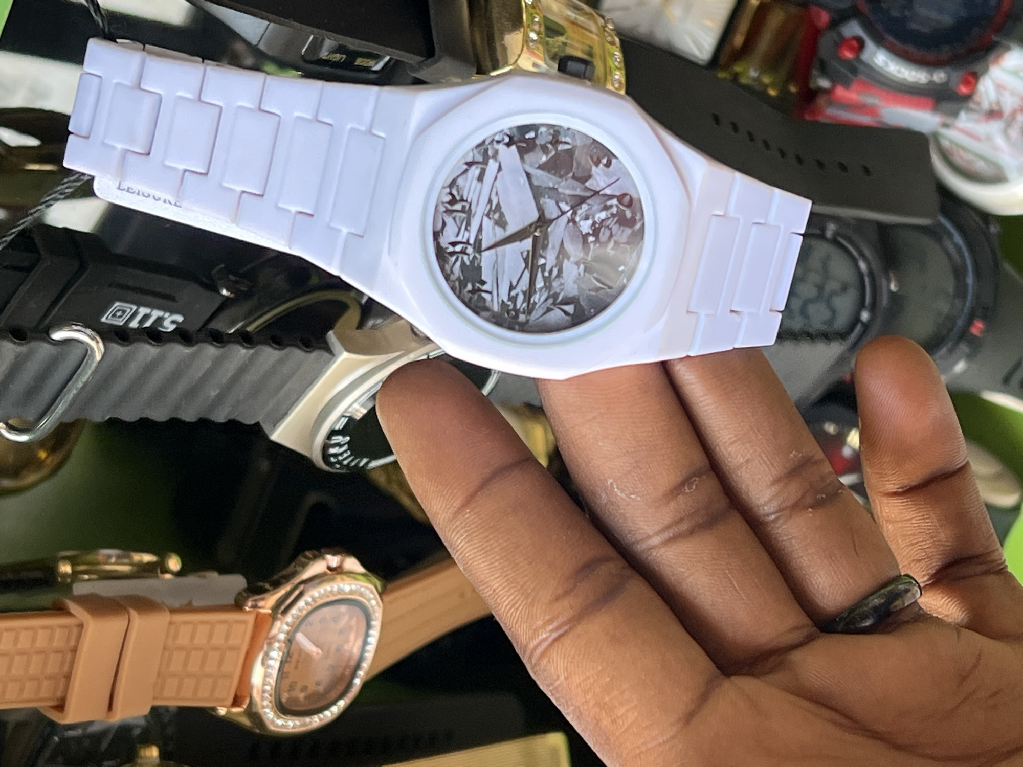 In Bukuru, Nigeria, an image captures a close-up of a person's hand holding a white wristwatch with a textured dial. Several other watches are visible in the background, including a gold-toned watch with a tan strap and a black digital watch. The scene appears to be an indoor setting, possibly a market or a shop, given the variety of items displayed.