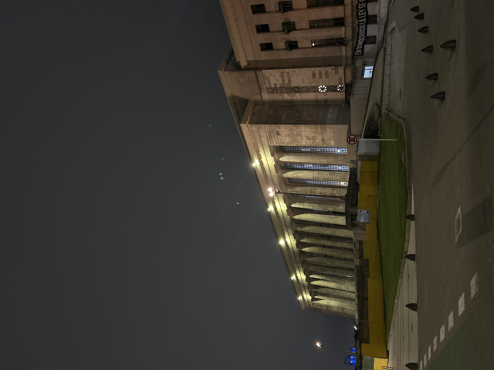 The image shows the Palace of the Argentine National Congress (Palacio del Congreso Nacional Argentino) at night, located in Buenos Aires, Argentina. The building is illuminated by lights lining its facade and columns. A patch of green grass is visible along the side of the building, bordered by a yellow fence. A sidewalk runs alongside the grass, marked with white squares and studded with conical bollards. A streetlamp casts a blue light further down the street. A sign hanging from the building reads "En Democracia la ley se cumple", which translates to "In democracy the law is obeyed."