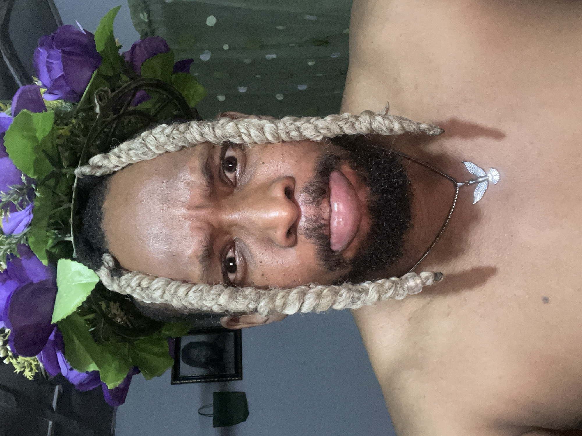 A portrait of a man facing the camera with his head tilted to the right. The man has dark skin, a beard, and light-colored braided hair framing his face. He is wearing a floral crown of purple flowers and green leaves. He is sticking his tongue out slightly. A silver necklace with a pendant is visible around his neck. The background is blurred, but a framed picture and a small, dark green object are discernible on a wall behind him. The image was not taken in Jos, Nigeria.