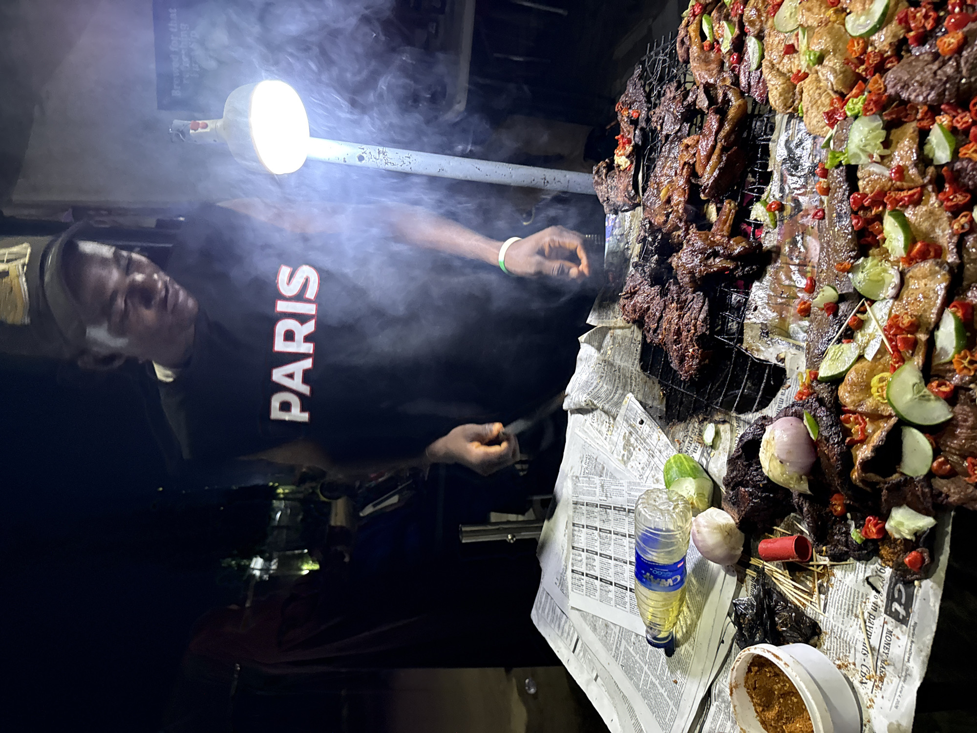 The image depicts an outdoor night market scene in Abuja, Nigeria. A young man wearing a black t-shirt with "PARIS" emblazoned across it stands behind a grill laden with grilled meat. He is looking down at the food, with a bent posture. Smoke billows around him from the grill, illuminated by a bright, overhead lamp. The grill itself is placed on top of a pile of newspapers, which also serve as a base for various food items and condiments.
In the foreground, a close-up view reveals a variety of grilled meats, some glistening and dark, others appearing more reddish-brown. The meats are garnished with slices of lime, small red chilies, and what appear to be pieces of cucumber. Scattered amongst the meat are whole garlic bulbs and onions. A plastic bottle filled with yellow liquid, likely cooking oil or a beverage, lies on its side on the newspapers. Next to it is a small white bowl containing a reddish-brown spice blend. Several wooden skewers and a red tube are also visible on the surface.
The background is dark, suggesting nighttime. Other figures are dimly visible in the background, including a person in a reddish garment and others further back, indicating the presence of other vendors or customers at the market. The environment appears to be a street or informal market setting, common in many parts of Abuja.