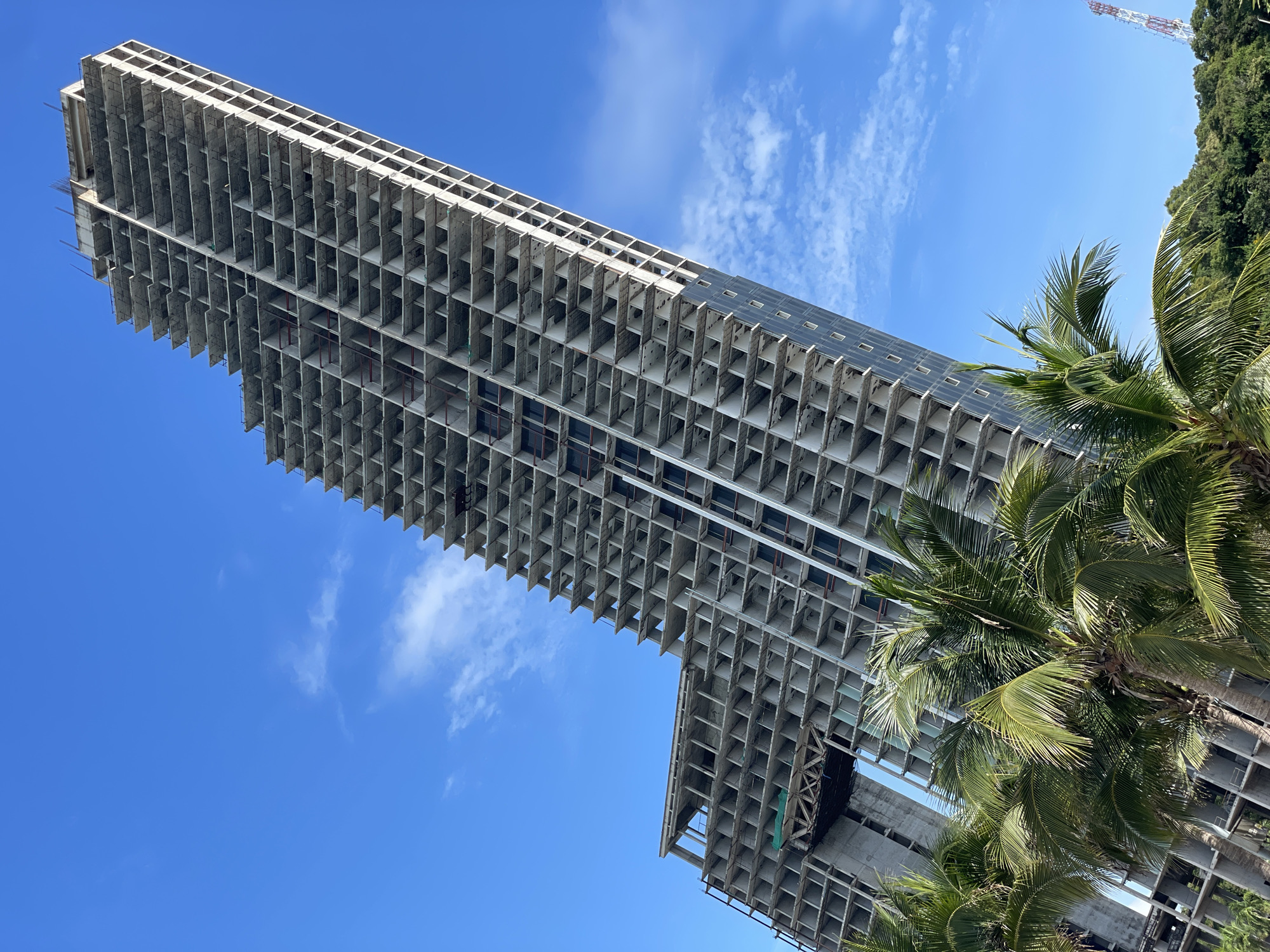 This image shows an unfinished skyscraper under construction in Pattaya City, Thailand. The building is tall and has a stepped design with many levels. The exterior is made of concrete, and the windows are not yet installed. Palm trees are in the foreground, and a lush green hillside is visible in the background. The sky is blue with scattered white clouds. The image appears to be taken from a low angle, looking up at the building.