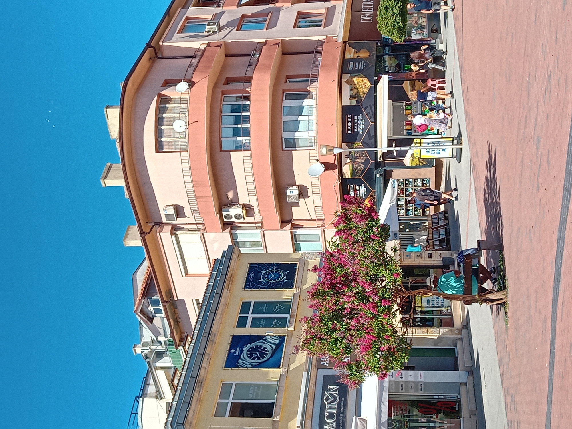 This image captures a street scene in Varna, Bulgaria, specifically on "Tsarigradska" street, a bustling commercial area. The scene is dominated by buildings with storefronts and apartments, bathed in bright daylight under a clear blue sky.
On the left, a yellow building features large windows displaying advertisements for watches, with prominent brands like "Aristo" and "Tissot" visible. Below, a storefront with a white awning displays a sign that reads "ACTION Watches & Jewelry". A vibrant pink-flowered tree stands in front of this building, adding a splash of color to the urban landscape.
To the right, a larger, salmon-colored building with rounded balconies and multiple windows occupies the center and right side of the frame. This building also houses shops on its ground floor, with awnings and signs indicating businesses. One sign prominently features "BUSHMILLS" and the text "BLACK BUSH", with an image of a castle. Another sign reads "DEMETRA".
The street is paved with red brick-like material. Several people are present in the scene. On the left, near the tree, a person wearing a teal shirt sits on a bench, looking at a phone. In the mid-ground, a group of people are walking and interacting. Further to the right, a few more individuals are seen walking along the street, some carrying bags. The overall atmosphere suggests a lively pedestrian area on a sunny day, with commercial activity and people enjoying the outdoor space.