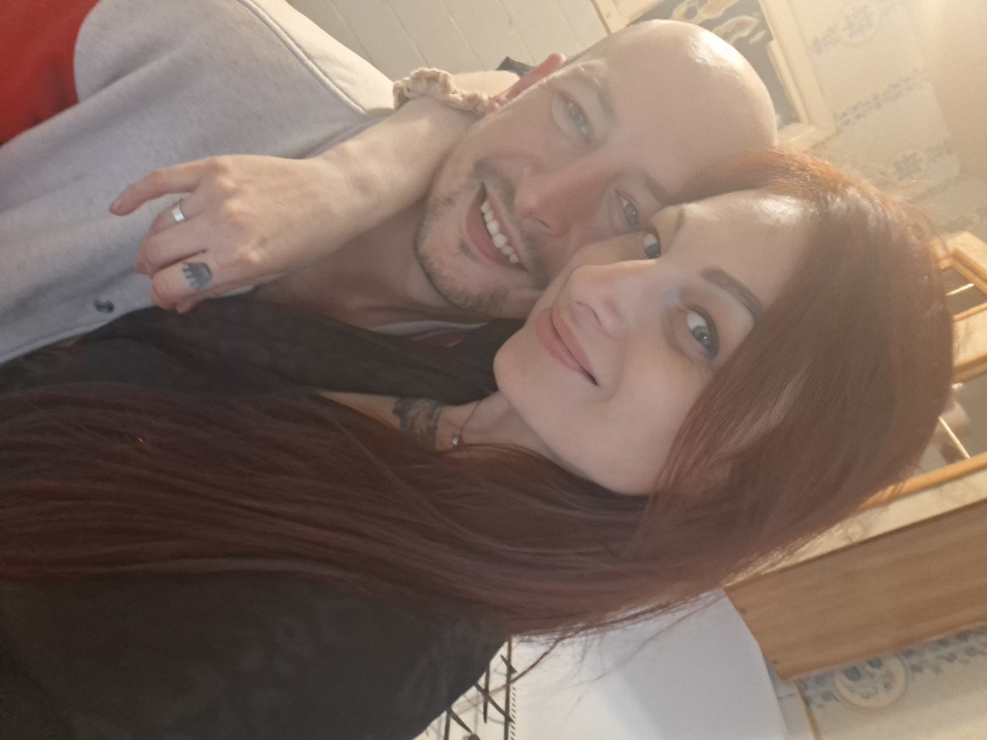 Two people, a man and a woman, are posing for a selfie. The man is bald and has a beard, and the woman has long, reddish-brown hair. They are both smiling, and the woman has her arm around the man's shoulders. The background appears to be a domestic interior, possibly a kitchen, with tiled walls and wooden accents. The lighting is warm, suggesting an indoor setting. There are no identifiable landmarks or features that would indicate the specific location within Забалоцкі сельскі Савет, Belarus.