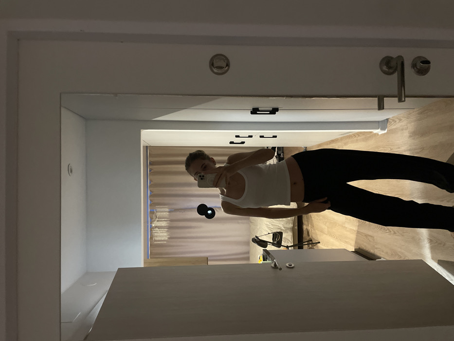 A woman in a white tank top and black leggings takes a selfie in a mirror. A desk or countertop reflects in the lower portion of the mirror. Behind her, a curtained window is visible. The room features a white doorframe and wall, with silver hardware (faucet, knobs) visible above. The mirror is positioned in a bathroom.