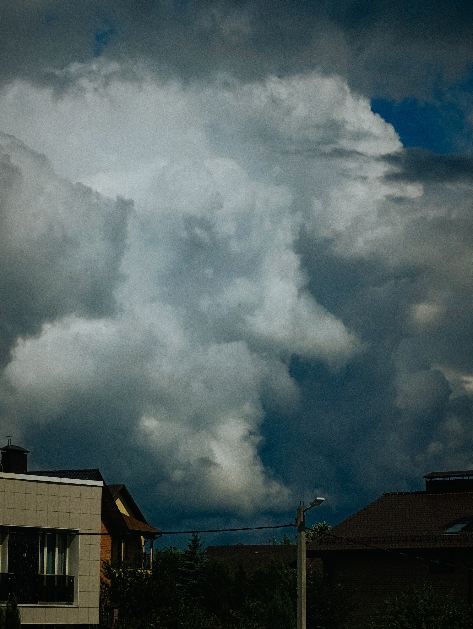 The image depicts a dramatic sky filled with dark, heavy storm clouds, casting a somber mood over a residential area. In the foreground, the rooftops of several houses are visible, interspersed with lush green trees. A modern, light-colored building with a large window stands to the left, while a traditional brick house with a dark roof is situated to its right. A streetlamp on a pole is also present in the scene. The overall impression is one of an impending storm in a suburban or semi-urban environment. The location is identified as Minsk, Belarus.