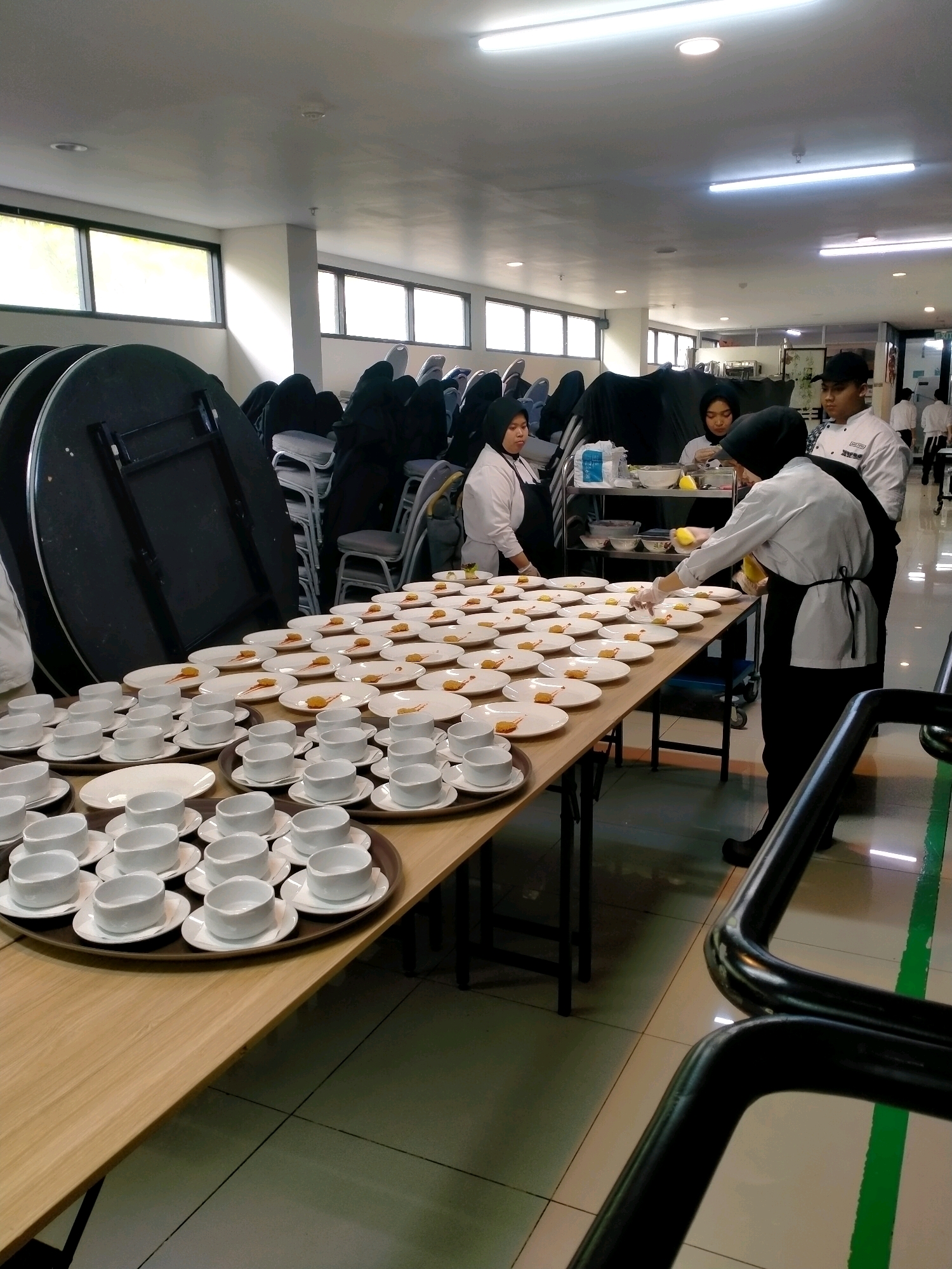 In a catering preparation area, likely within a convention center or hotel banquet facility in Batu, Indonesia, a long, wooden table is laden with prepared food items and servingware. The table, positioned in the foreground, is covered with rows of small white plates, each holding a portion of food, possibly an appetizer or dessert, with a drizzle of sauce. Surrounding these plates are numerous stacks of white teacups and saucers, arranged on trays.
Several individuals, dressed in chef's uniforms, are actively involved in the preparation. In the center of the image, two women in chef attire, one wearing a hijab, are working. One is holding a yellow object, possibly a piece of fruit or a garnish, and appears to be plating food. Another person, a young man in a chef's uniform, stands behind them, observing. To the left, another woman in a chef's uniform is also engaged in the setup. In the background, more individuals in chef attire are visible, further down the hall.
The environment suggests a functional, clean setting. The walls are light-colored, and fluorescent lighting illuminates the space. Large windows are visible in the background, letting in natural light. Stacked round tables, covered with dark fabric, and chairs are neatly arranged against a wall, indicating a space that can be reconfigured for events. The floor is tiled, with distinct green lines marking pathways or areas. The overall scene depicts a professional catering operation in progress.