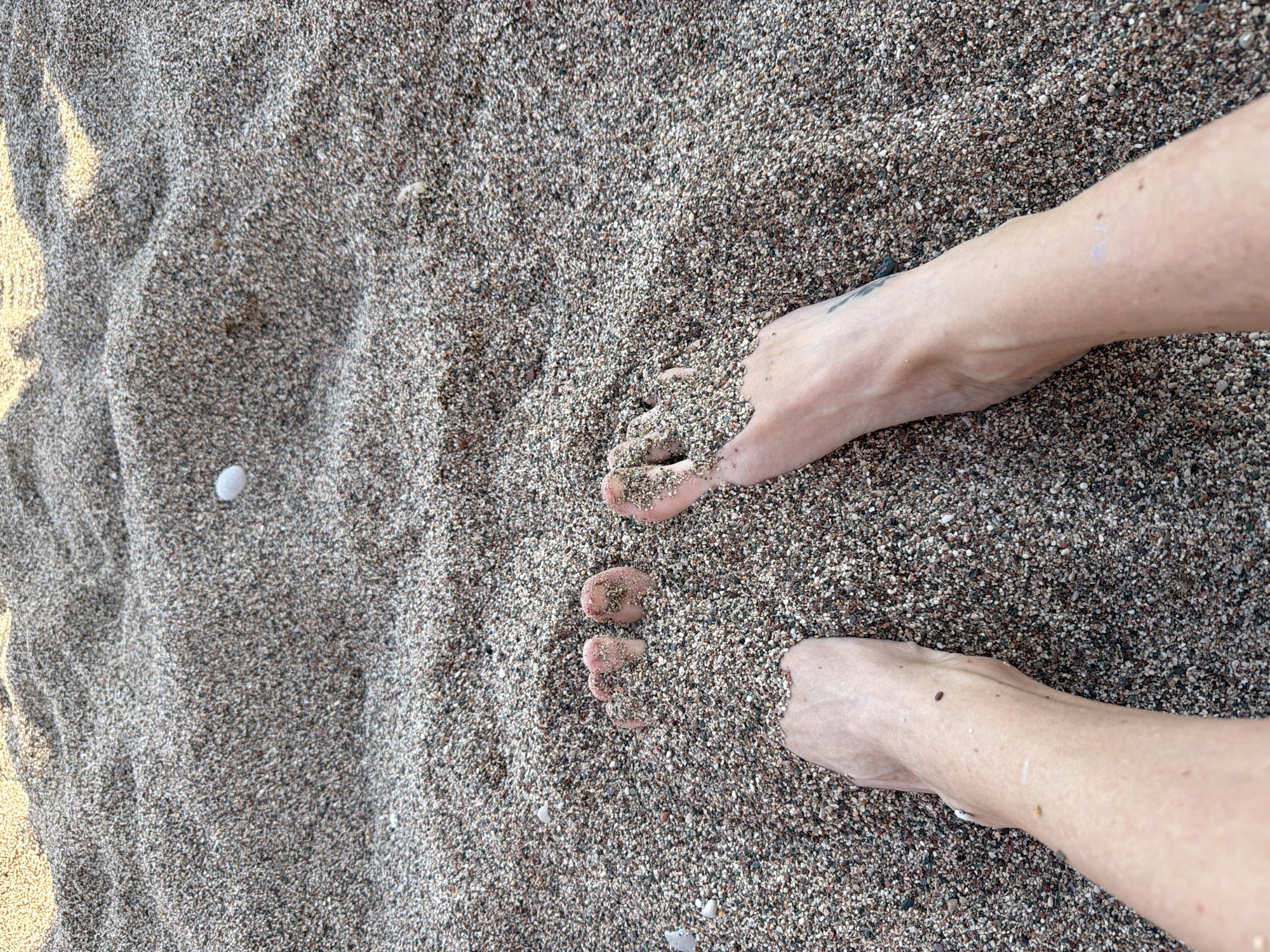 The image shows a pair of feet on a sandy beach. The sand appears to be coarse and gray. The feet are fair-skinned, with sand covering the toes. There is a light-colored pebble or shell near the left foot. The specific location (city/country) is not discernible from the image.