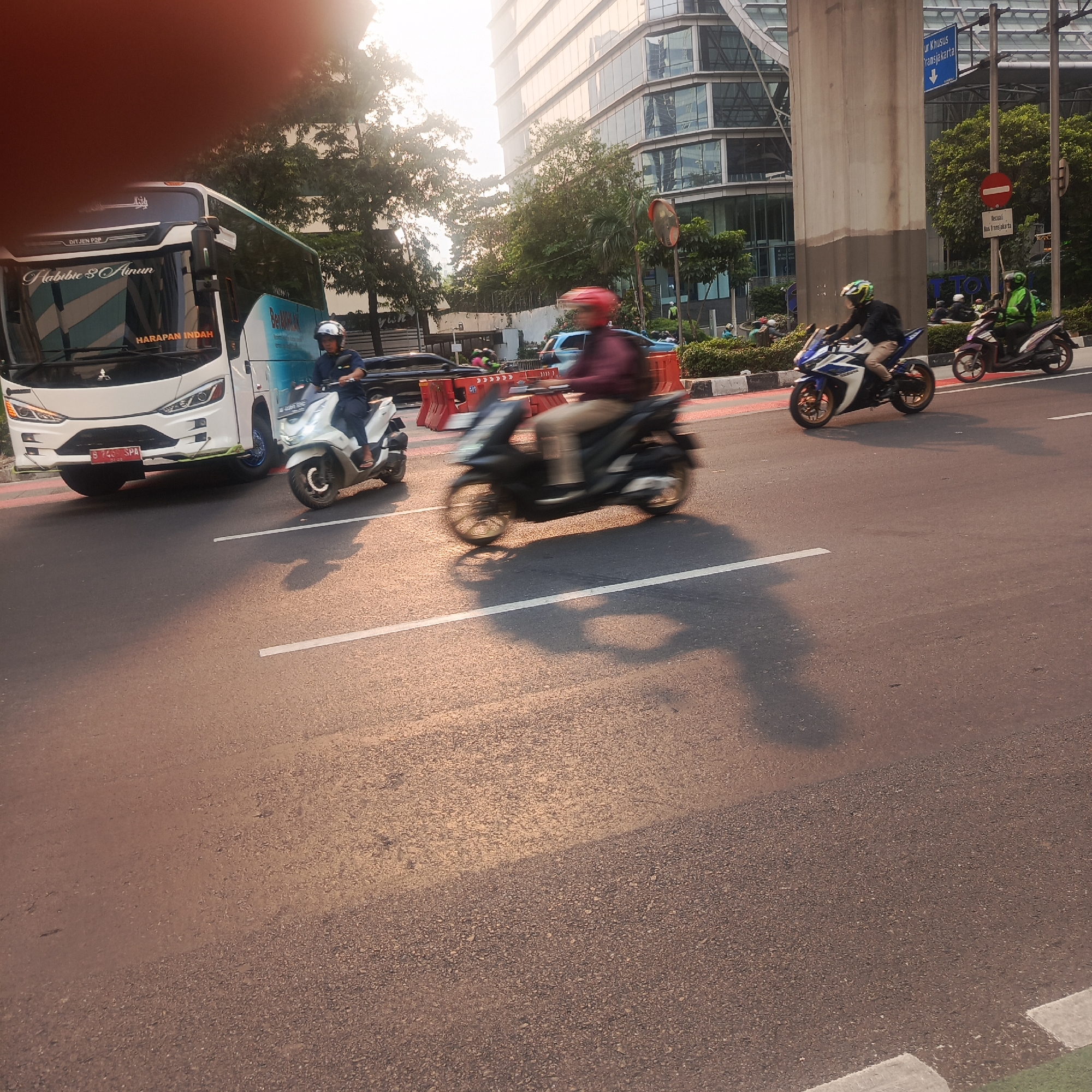 This is a photograph taken from a low angle on a sunny day, likely in the afternoon, depicting a street scene in South Jakarta, Indonesia, possibly along Jalan Sudirman or a similar major thoroughfare due to the modern high-rise buildings in the background and the presence of public transportation signs. The foreground is dominated by an asphalt road with white lane markings. The lighting suggests the sun is low in the sky, casting long shadows.
In the middle ground, several vehicles are visible, some in motion and others stationary. On the left, a white bus with blue and teal accents is partially visible. It has text on its front, including "HABIBIE & AINUN" and "HARAPAN INDAH". A white scooter is in front of the bus, with a person wearing a dark uniform and a helmet riding it.
Moving towards the center and right, a person on a scooter with a red helmet is captured in motion blur, suggesting rapid movement. Further to the right, another motorcycle, a sports bike with blue and white fairings and gold rims, is also in motion blur, ridden by a person wearing a helmet. Another scooter, ridden by a person in a green jacket, is visible behind it. Orange traffic barriers are also present on the side of the road.
The background features tall, modern glass-fronted buildings, characteristic of a central business district. Trees line the median and sides of the road. A traffic sign with blue lettering and a down arrow, possibly indicating a bus lane or route, is visible on a tall pole. Another traffic sign with a red circle and horizontal bar is also present. The overall atmosphere is one of urban activity and traffic.