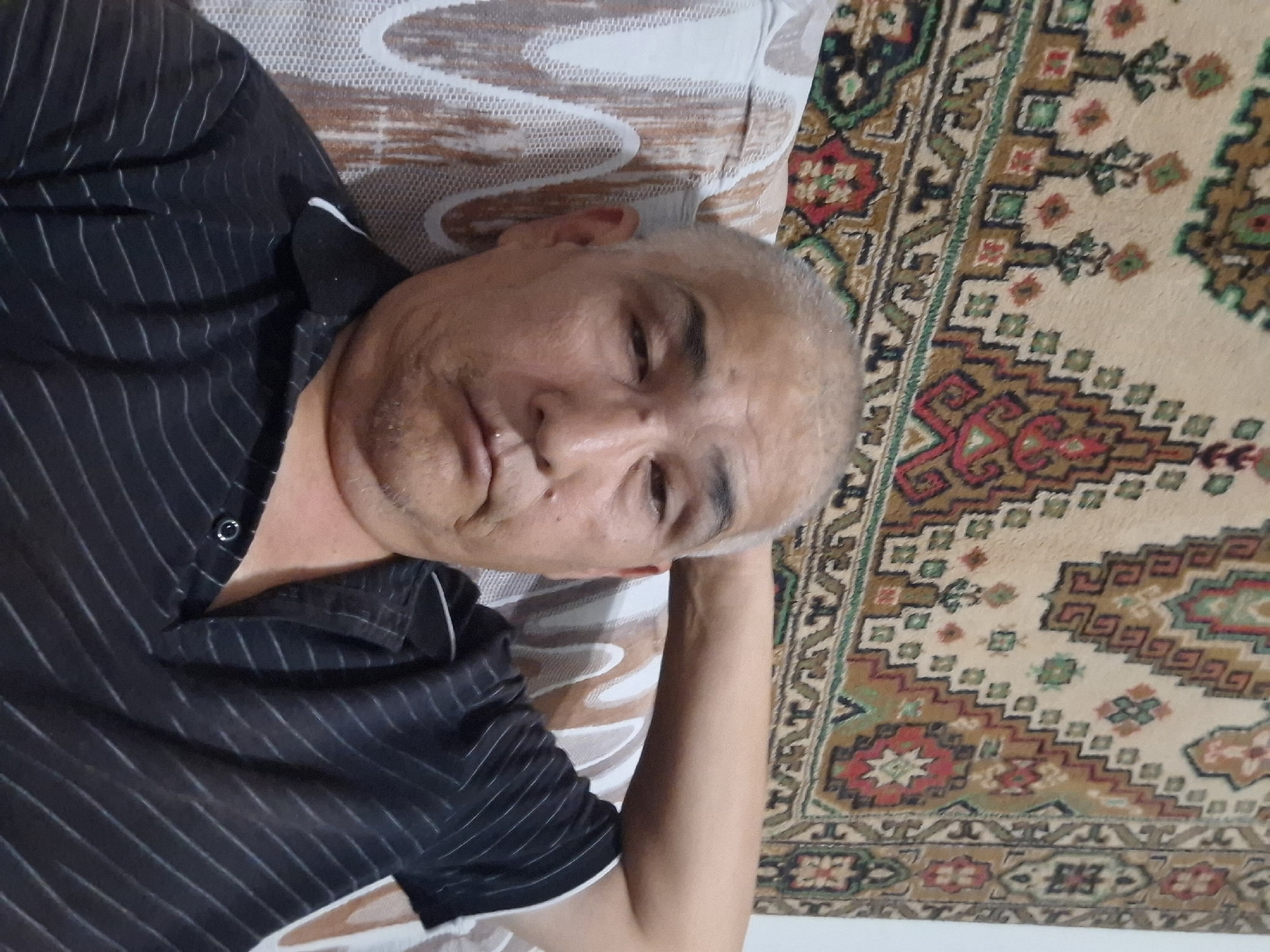 A close-up, overhead shot shows a middle-aged East Asian man resting his head on his arm, looking directly at the camera. He has short, graying hair, a light beard, and is wearing a dark, striped polo shirt. He appears to be indoors, seated on a couch with a patterned rug behind him. The rug features intricate geometric designs in red, green, brown, and beige. The couch has a white, textured fabric with a brown stripe pattern. The lighting suggests a natural light source from the right. The man's expression is neutral. The scene appears to be in a domestic setting in Almaty, Kazakhstan, as indicated by the traditional rug and interior decor.