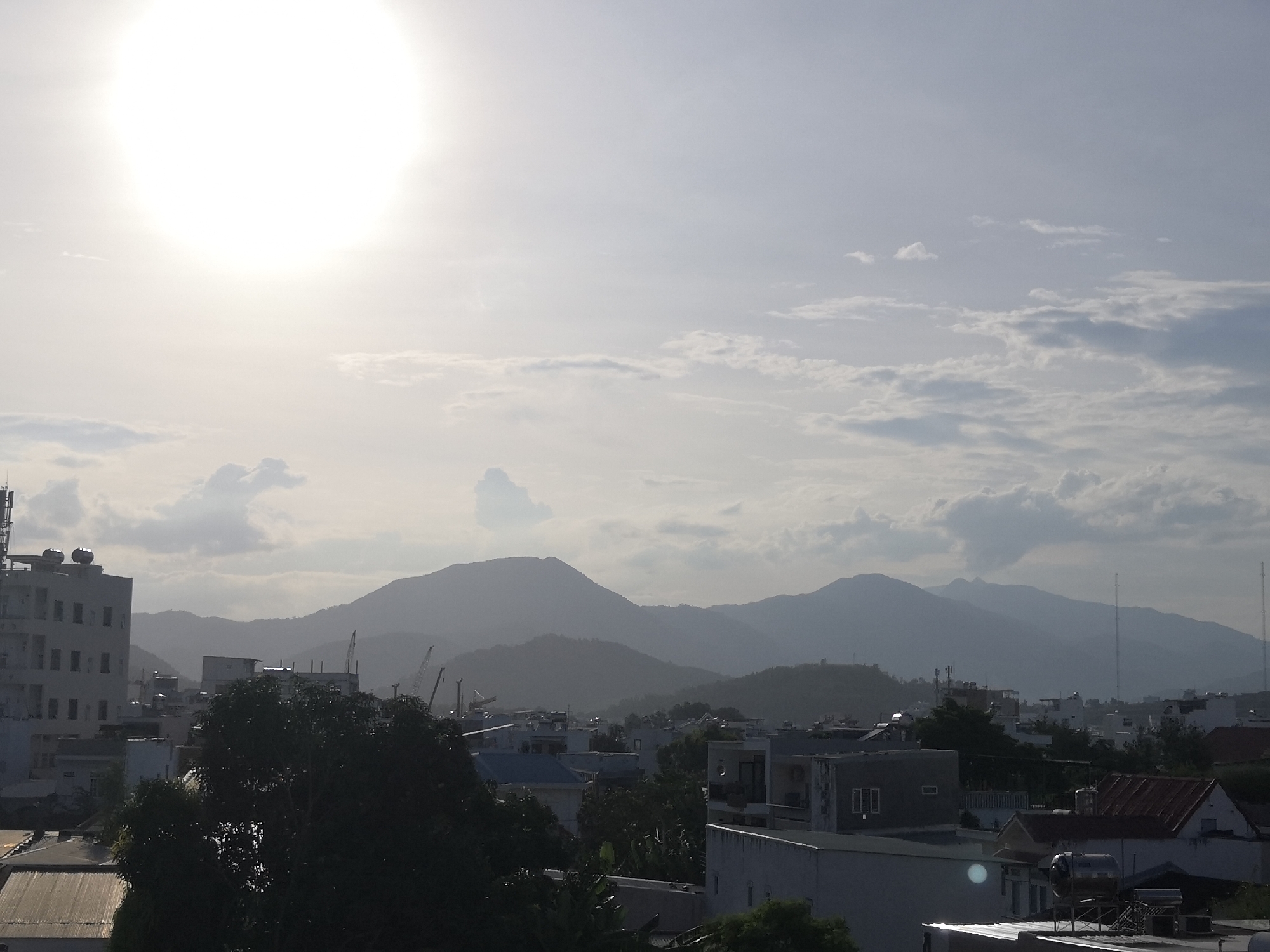 The image captures a hazy, high-angle view of a city with mountains in the background, likely in Bac Nha Trang Ward, Vietnam. The sun is prominently featured in the upper left quadrant, casting a bright glare that washes out the sky. Silhouetted against the hazy sky are several mountain ranges. In the foreground and midground, a dense urban landscape is visible, characterized by a mix of buildings, rooftops, and lush green trees.
Several buildings are identifiable: a tall, multi-story building with visible windows and what appear to be solar water heaters on its roof stands on the left. Scattered throughout the city are smaller residential and commercial structures, some with pitched roofs, others with flat roofs. Construction cranes are visible among the buildings, suggesting ongoing development. Several antennas and communication towers are also present. The overall impression is of a densely populated urban area nestled near a natural mountainous landscape, under a bright but diffused sun. There are no people visible in the image.