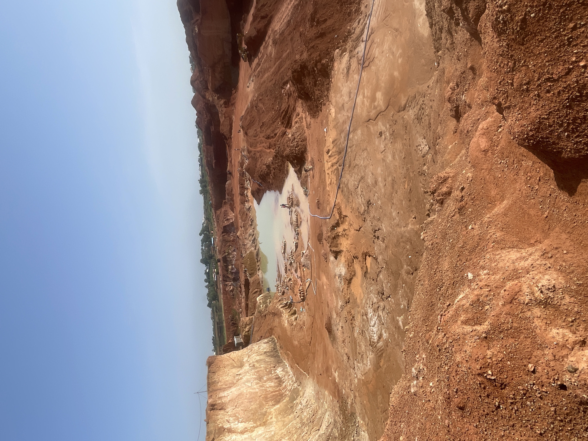 The image depicts an open pit mine in a rural area near the city of Jos, Nigeria. The environment is arid and characterized by reddish-brown soil and rock formations, indicating the presence of iron oxide. A pool of stagnant water is situated in the center of the mining pit. Several individuals are present in the mine, seemingly engaged in the extraction of minerals or other resources. Blue piping runs across the area. In the background, a settlement of houses is visible among trees. The sky is pale blue, suggesting a clear, sunny day.
