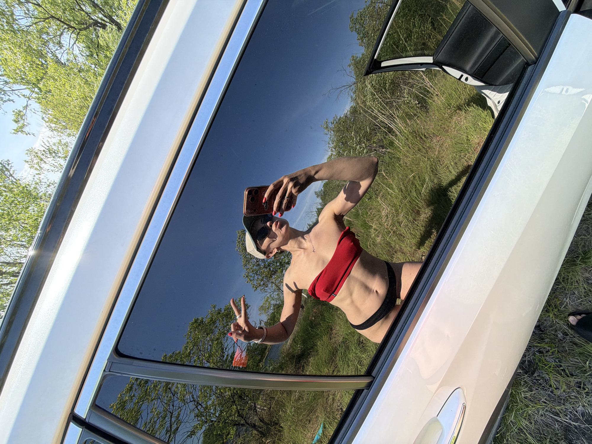 A reflection in the dark window of a white car reveals a person in a red top and black bottoms, holding a phone and making a peace sign. The car is parked in a grassy area with trees in the background. Раздольное, Russia is not explicitly identifiable from the image.