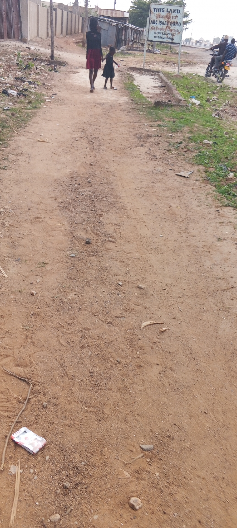 A medium shot depicts a dirt road in Keffi, Nigeria. Two individuals, an adult female and a young girl, are walking hand-in-hand along the road. A sign on the right side of the road indicates "THIS LAND BELONGS TO ARC ISAA GONO" along with contact information. A motorcycle with two occupants is visible on the right side of the frame. In the foreground, there is discarded trash on the ground. Buildings and fences are visible in the background. The environment appears to be a residential or developing area.