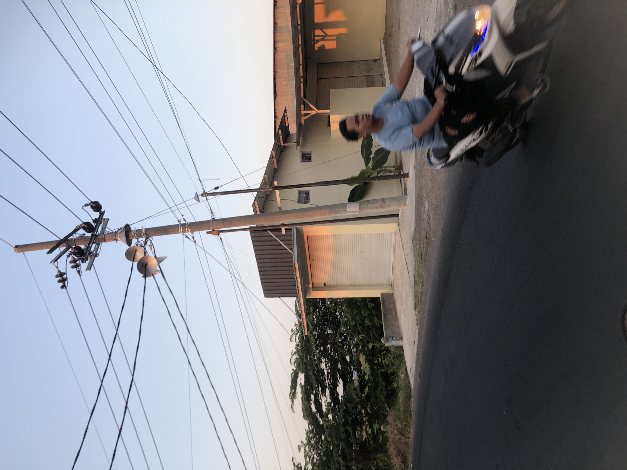 A person rides a motorcycle on a paved road in Glagah Arum, Indonesia, with utility poles and power lines overhead. The person, wearing a light blue t-shirt and dark pants, is in motion and their face is blurred. The motorcycle is white with some black and blue accents. To the right of the road is a light-colored building with a corrugated metal roof and a large garage door. In the background, there are dense green trees and vegetation. The sky is a pale blue, indicating either early morning or late afternoon. The scene is captured with a shallow depth of field, focusing on the motorcycle and rider.