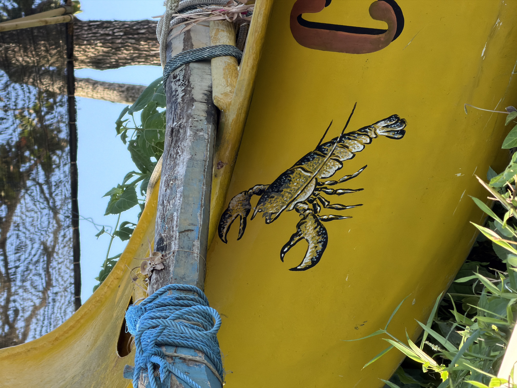 A close-up shot depicts a section of a yellow boat or structure, likely located in North Kuta, Indonesia. A detailed painting of a lobster adorns the yellow surface. Adjacent to the yellow structure is a weathered wooden post, secured with blue rope. Green foliage and other structural elements are visible in the background. The sky peeks through the upper left corner of the frame. The composition suggests an outdoor setting.
