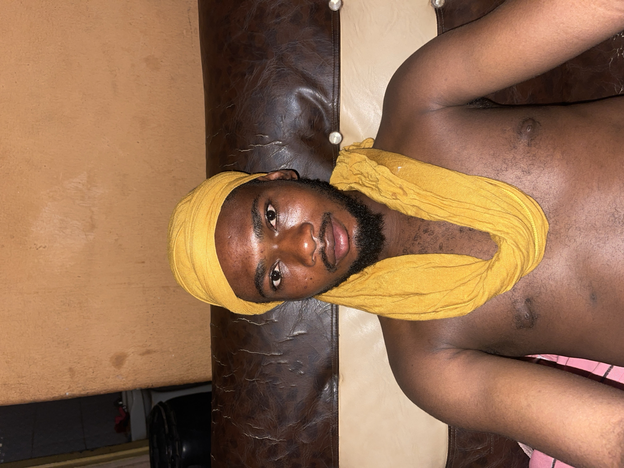 A man with dark skin, a beard, and a yellow head covering is lying on a brown and beige surface. He is shirtless. The location appears to be indoors.