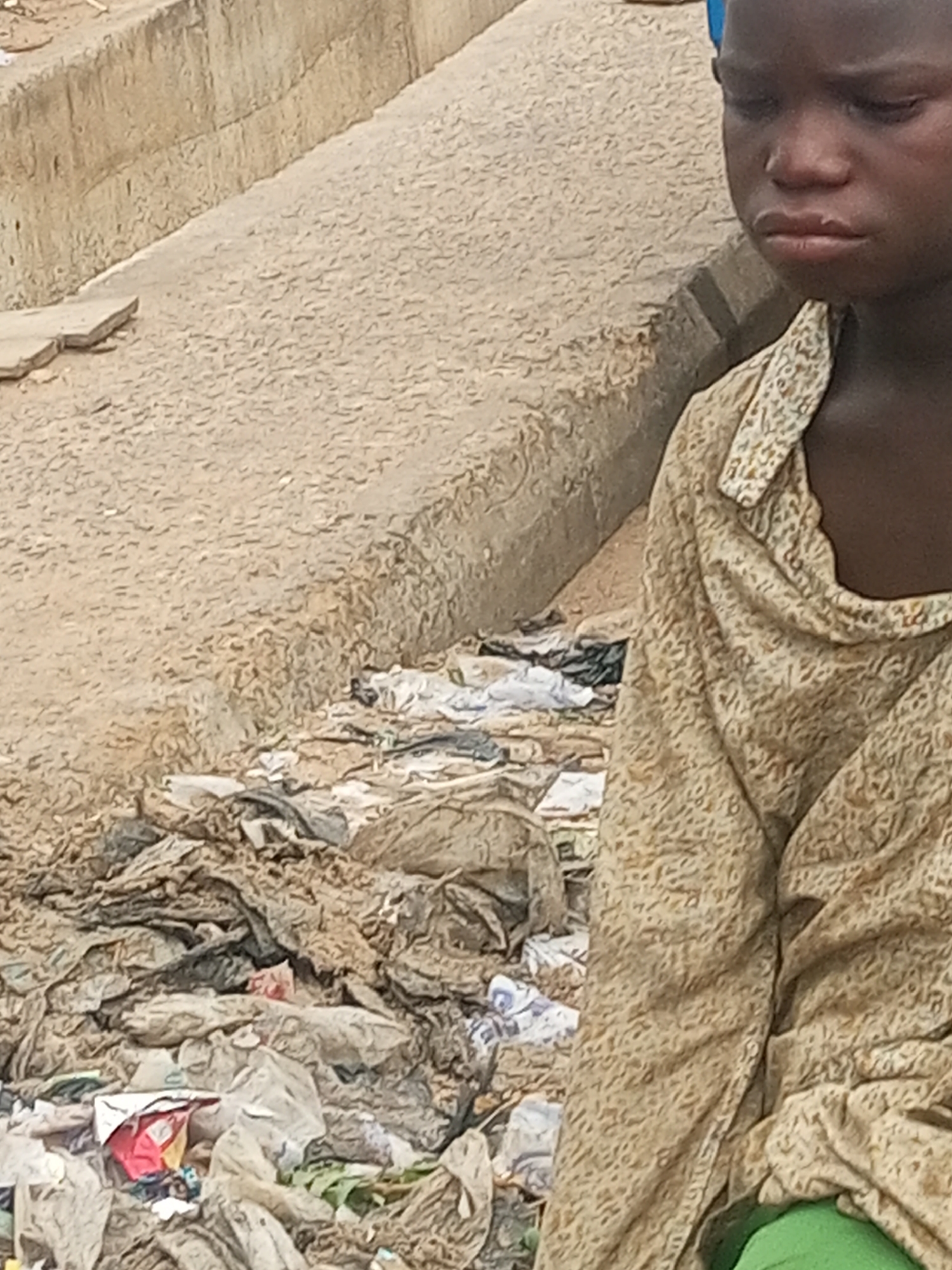 The scene depicts a young individual, likely a child, positioned on the right side of the frame, alongside a concrete structure and an accumulation of refuse. The location is the city of Unknown, Unknown.
The child, with dark skin, is partially visible from the forehead to the chin, facing slightly away from the camera with a downward gaze. Their eyes are downcast, and the lips are slightly pursed. They are wearing a light beige garment with a small, repetitive organic pattern, featuring a visible collar. A bright green fabric is discernible beneath this garment on the lower right.
To the left and center of the frame, a textured, light brown ground surface is present, appearing to be packed earth or rough concrete. A concrete barrier or wall traverses diagonally from the upper left toward the center, exhibiting cracks and a coarse texture. Below this barrier, a substantial pile of discarded materials, primarily composed of plastic bags, wrappers, and unidentified debris, is observed. The refuse is densely packed and varied in color, dominated by muted tones of gray, white, and brown, with minor accents of red and green from packaging. A flat, broken concrete slab is visible on the upper left ground surface. The lighting is consistent with outdoor daylight conditions.