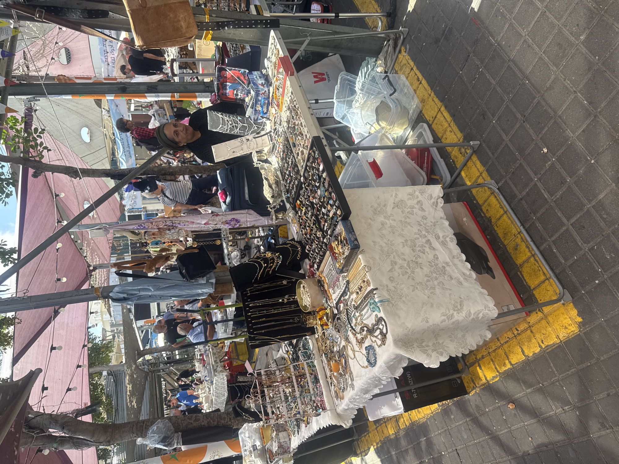 This image shows an outdoor market stall in Tel Aviv, Israel. An assortment of jewelry, including rings, necklaces, and bracelets, is displayed on a table covered with a white floral tablecloth. A woman is standing behind the table. Behind the stall are clear plastic storage containers. Overhead, there are awnings and string lights suspended above the stalls. The ground is paved with gray bricks, with a yellow painted curb. Signs with Hebrew and Arabic script are visible.
