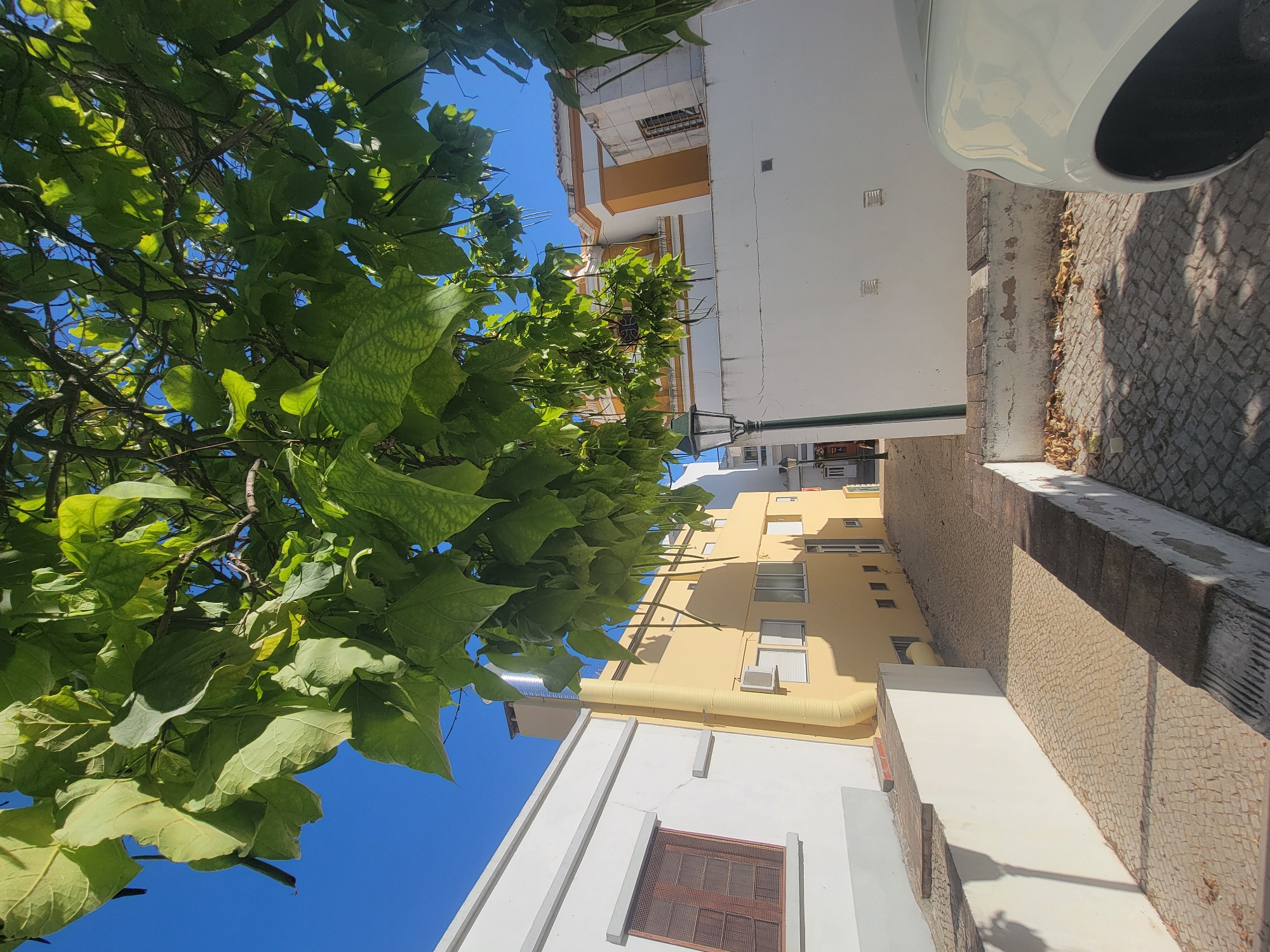 The image captures a sun-drenched, narrow street in Faro, Portugal, with a prominent tree canopy in the foreground casting dappled shadows. To the left, a white building with a dark-framed window with a metal grate is visible. Adjacent to it is a yellow building with multiple windows and an air conditioning unit, featuring a large, curved yellow pipe running vertically up its facade towards a metal chimney. The street itself is paved with small, irregular cobblestones. Further down the street, a white building with orange trim and windows can be seen. A dark green lamppost stands on the right side of the street. The right side of the image shows the partial view of a white car. The sky is a clear, vibrant blue. The scene appears to be in a historic part of the city, likely residential or commercial, given the architectural styles.