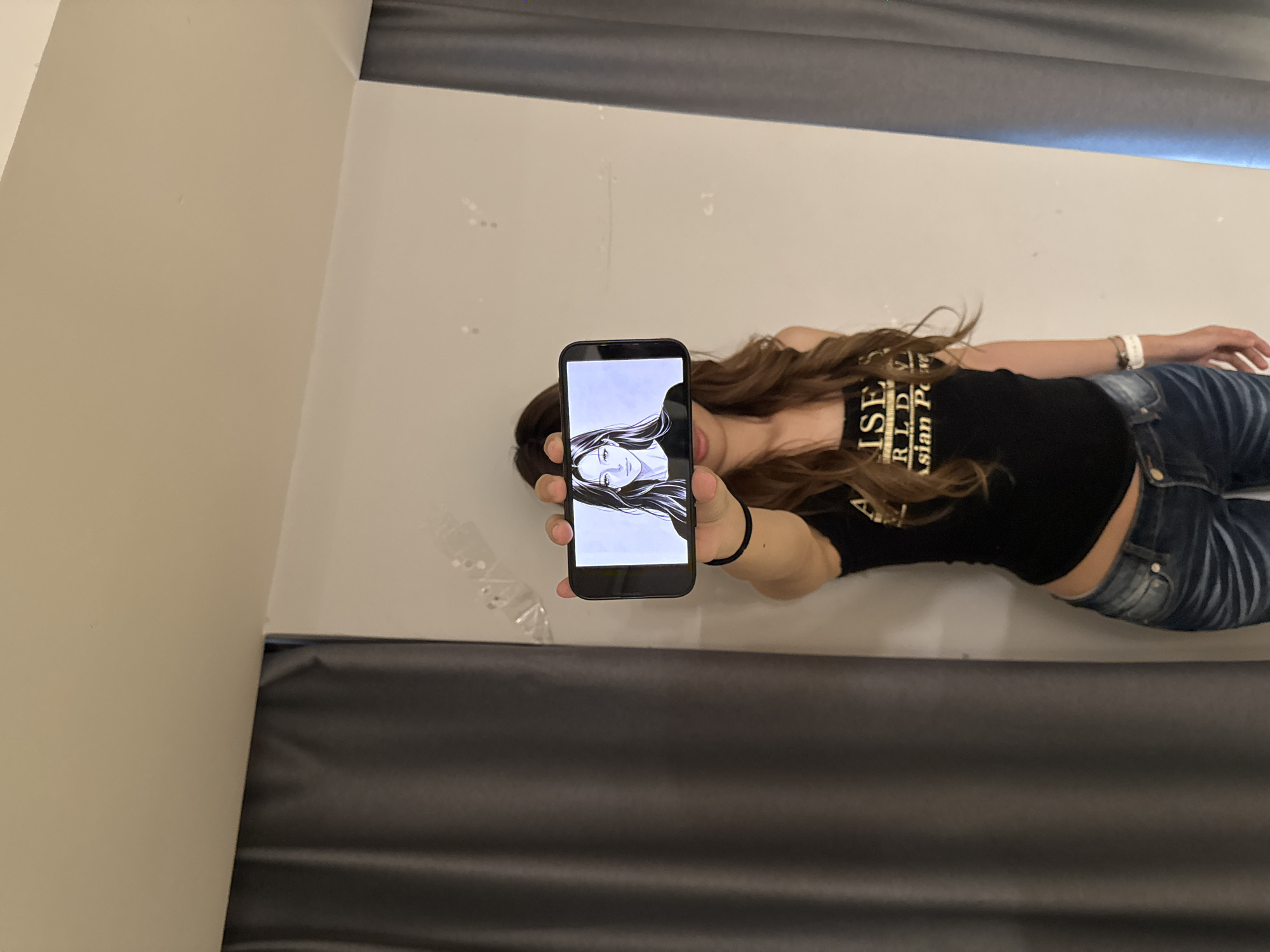 A woman is lying on a gray surface, possibly a bed or couch, holding a smartphone in her left hand. The screen of the phone displays a black and white portrait. The woman is wearing a black t-shirt with gold lettering and blue jeans. Her hair is long and brown. The background appears to be a white wall with a gray fabric or drape along the top and bottom edges. The location in the city of Liangjiang, China, cannot be determined from this image.