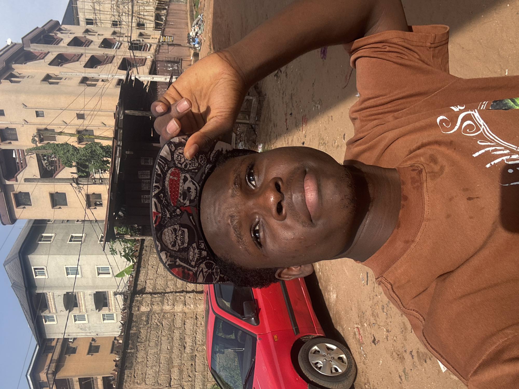 A dark-skinned man wearing a brown t-shirt and a skull-patterned baseball cap is holding the brim of the cap, looking directly at the camera. The background reveals a red car, a concrete wall, and several buildings. The overall location is Onitsha, Nigeria. The scene appears to be outdoors, with mixed terrain, including concrete and dirt. There is litter present in the background.
