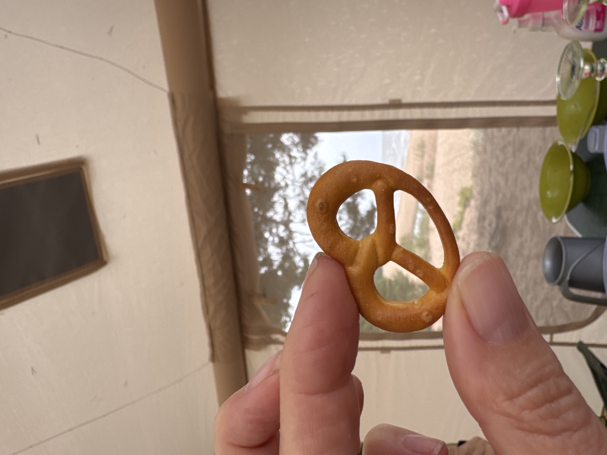 A close-up shot shows a hand holding a pretzel against a blurry background of a sandy beach with trees and the sea. The pretzel is golden brown with a classic knotted shape. The hand has visible fingernails and skin texture. In the background, a tan-colored tent interior is visible, with a window or opening revealing the outdoor scene. On a surface within the tent, there are green bowls, a metal cup, and a pink bottle. The overall impression is one of a casual outdoor setting, possibly a beach picnic or camping trip, with a focus on the snack. The location is specified as Ozerki (Krasnodolinskaya), Russia.