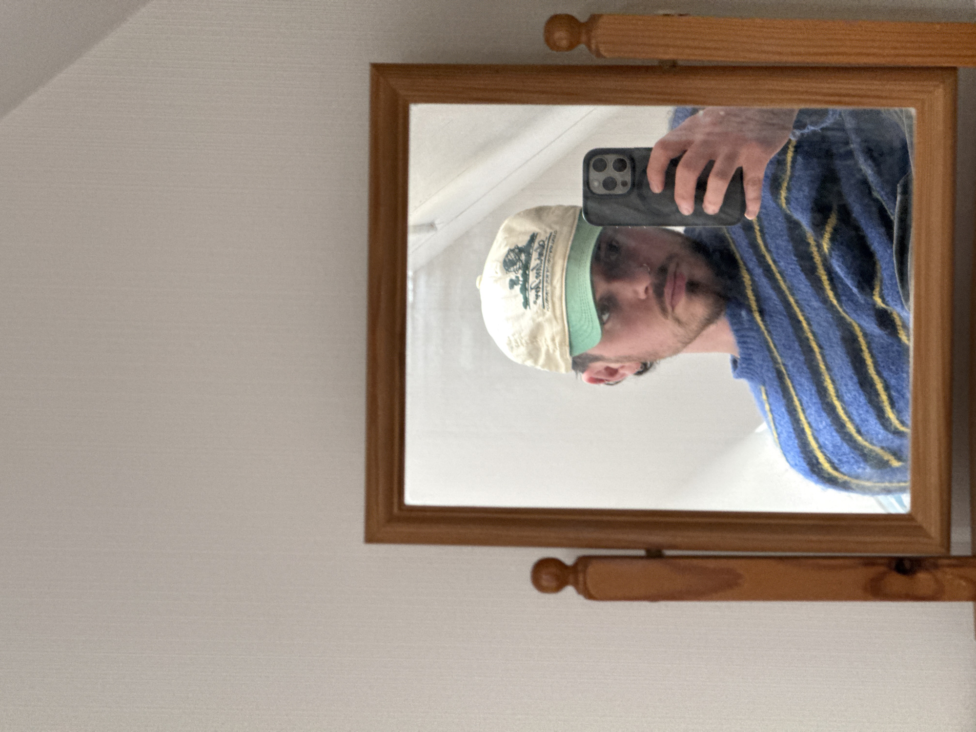 A man is taking a selfie in a wooden-framed mirror. The mirror is hanging on a white wall. The man is wearing a tan baseball cap with a green brim and a blue sweater with yellow stripes. The man is holding a smartphone in his hand. The location is Unknown, Unknown.