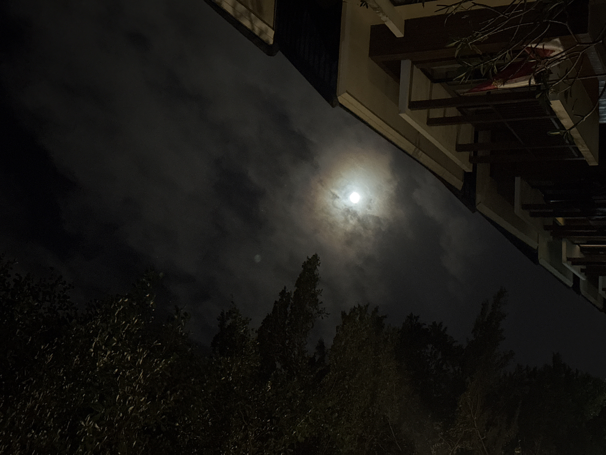 The image shows a nighttime scene in Kuta Selatan, Indonesia. A cloudy sky is visible with a bright full moon at the center. Part of a building with exposed wooden beams and a dark roof is on the right side of the image. Trees fill the lower portion. The scene is dark and lacks details due to the low light conditions. No people or actions are visible in the frame.