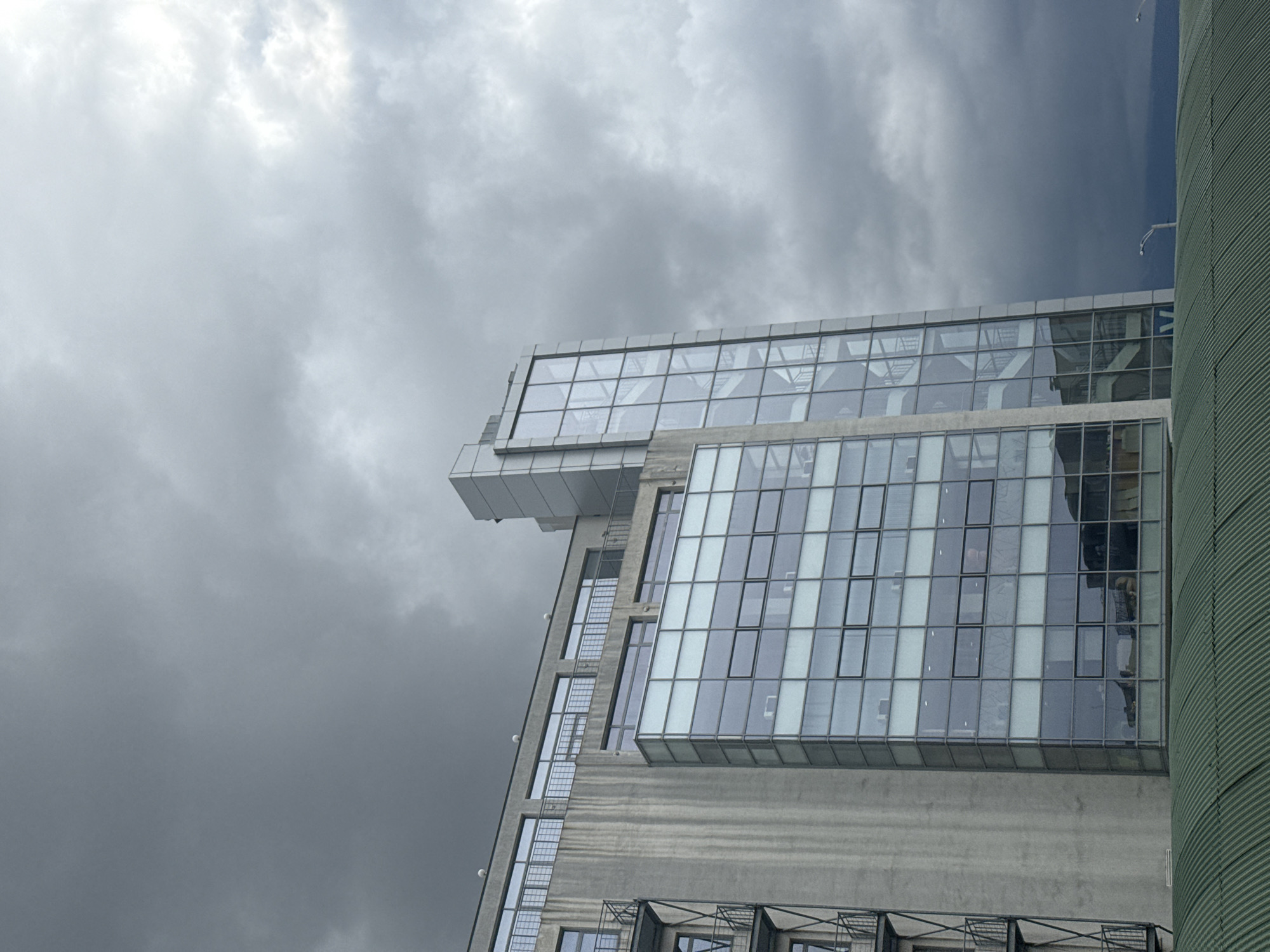 The image shows the architecture of a building against a cloudy sky. The building features a concrete structure with a prominent glass facade. The facade is composed of numerous rectangular glass panes. The clouds are predominantly gray. The building is the National Gallery for Foreign Art located in Sofia, Bulgaria. The architectural style appears modern. There are no discernible people or actions taking place in the immediate view. The overall environment appears urban. A green colored cylindrical structure is on the right.