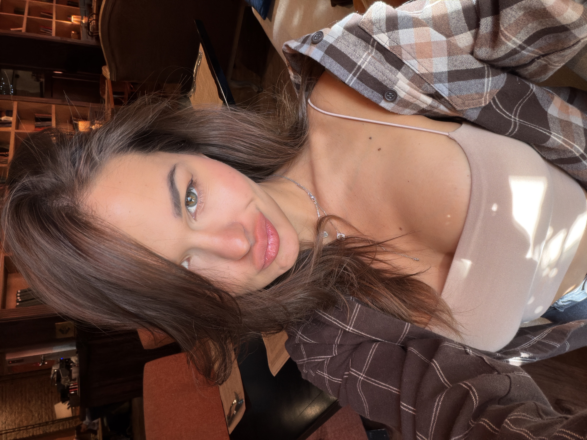 A woman with brown hair, wearing a beige top and a brown and white plaid shirt, is posing for a selfie. The background features wooden shelves and a chair, suggesting an indoor setting, possibly a cafe or library. The exact location in Krasnoyarsk, Russia, is not discernible from the image alone.
