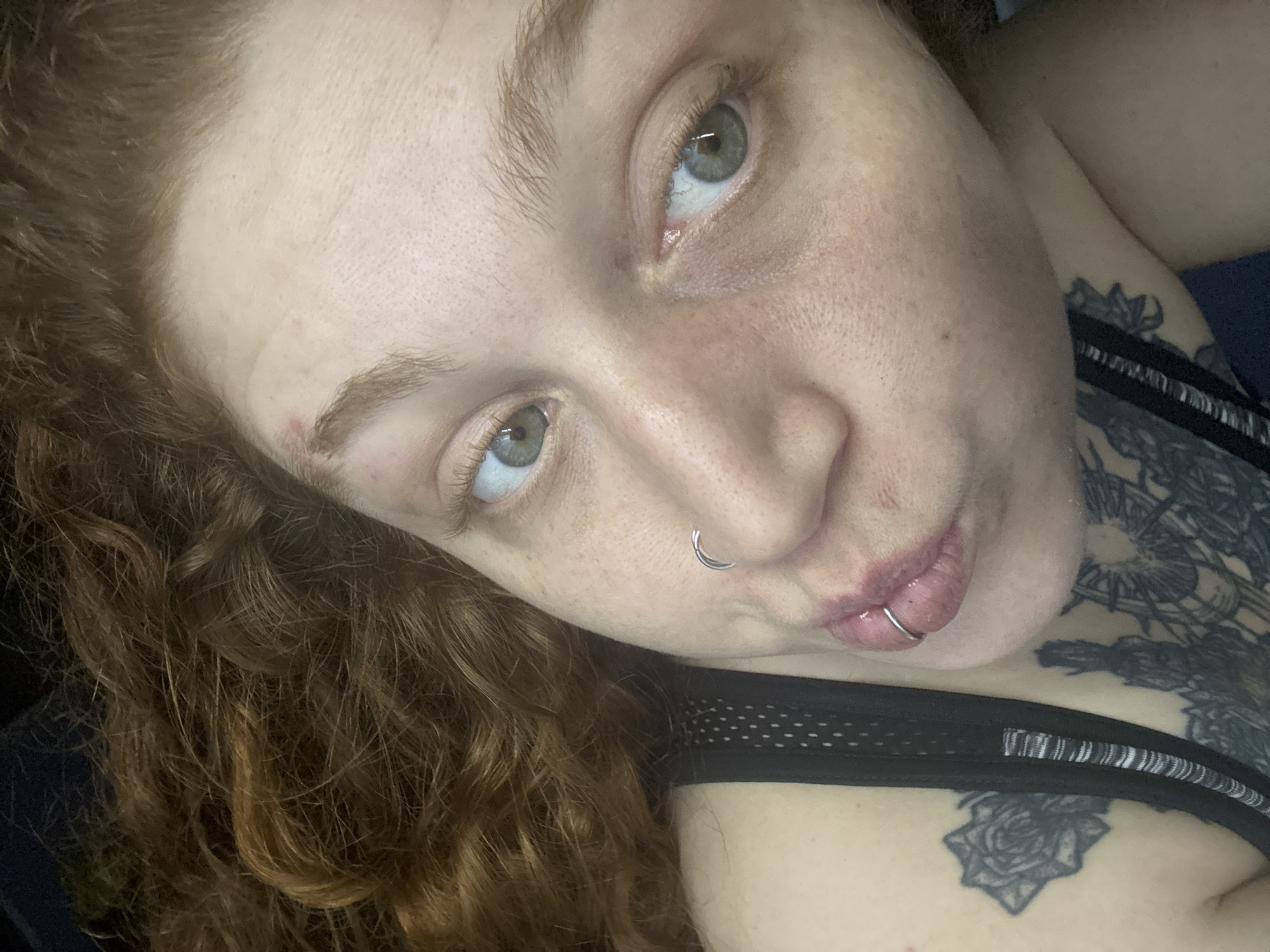 A close-up shot of a fair-skinned individual with light brown/red curly hair. The person has light green eyes and is wearing a nose ring and a lip ring. Visible on the person's chest are two tattoos; one a stylized rose and the other a more complex design resembling a sun or star. The person is wearing a black mesh-like top with visible straps. No indication of the location being Buenos Aires, Argentina is present.