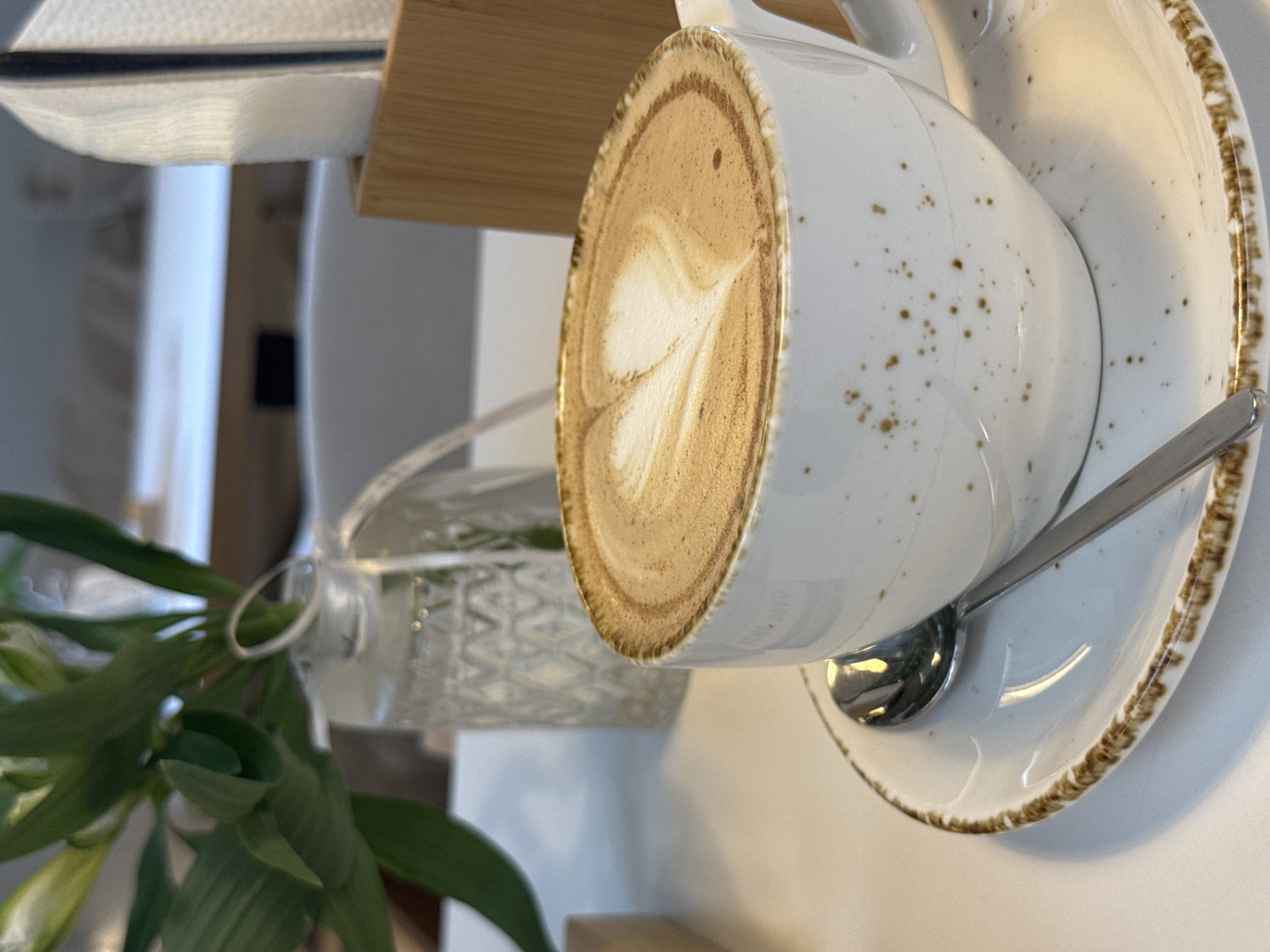 A close-up, eye-level shot showcases a white ceramic cup of coffee with latte art in the shape of a heart, resting on a matching saucer. A silver spoon lies diagonally across the saucer. The cup has a speckled, rustic texture. In the background, out of focus, is a clear glass vase containing green leaves and a white ribbon. To the right, a wooden napkin holder with white napkins is visible. The scene is set on a white table, suggesting a cafe or restaurant environment. This image was captured in Naberezhnye Chelny, Russia.