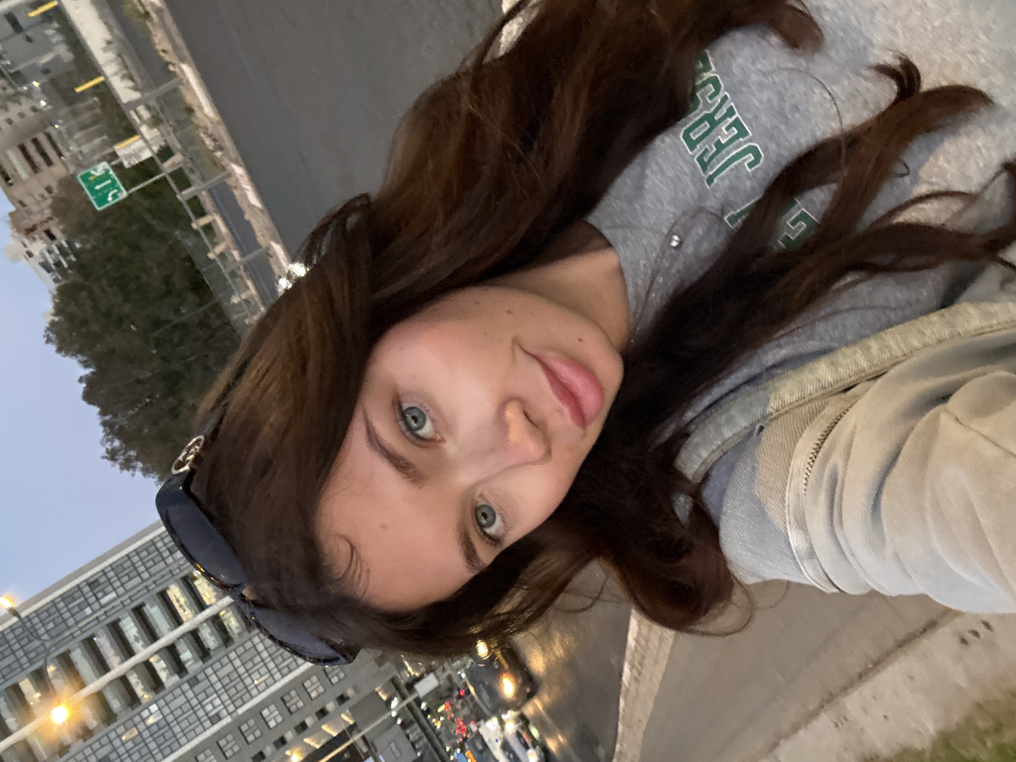 A selfie captured at street level shows a woman with long, dark brown hair and light blue eyes, wearing a gray hooded sweatshirt with green lettering that appears to spell "JERSEY." She is also wearing sunglasses, visible on the side of her head.
The background includes a building with multiple windows and a street with traffic lights. A green street sign is also visible. The scene appears to be in an urban setting, identifiable as Tel Aviv based on the street sign and the style of architecture of the buildings. It is evening, as indicated by the subdued lighting and the presence of streetlights.