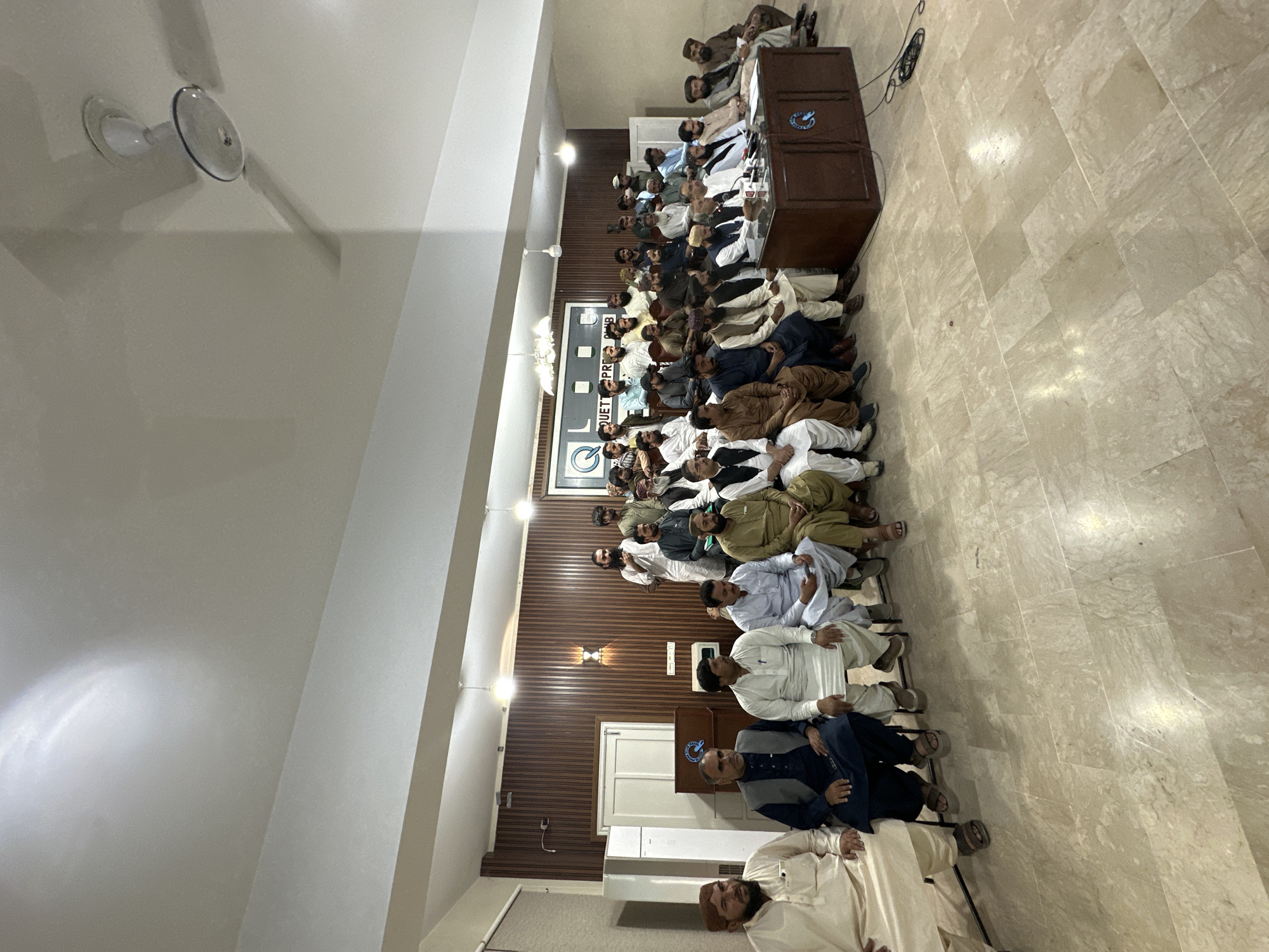 The image shows a room in Quetta City Tehsil, Pakistan, filled with people seated in rows. A dark-wooden podium stands at the front, equipped with microphones. A logo with the words "QUETTA PRESS CLUB" is displayed on a wall. The people present are predominantly men, dressed in traditional clothing. The room has white walls, a wooden-paneled section, and a tiled floor. A ceiling fan is visible.