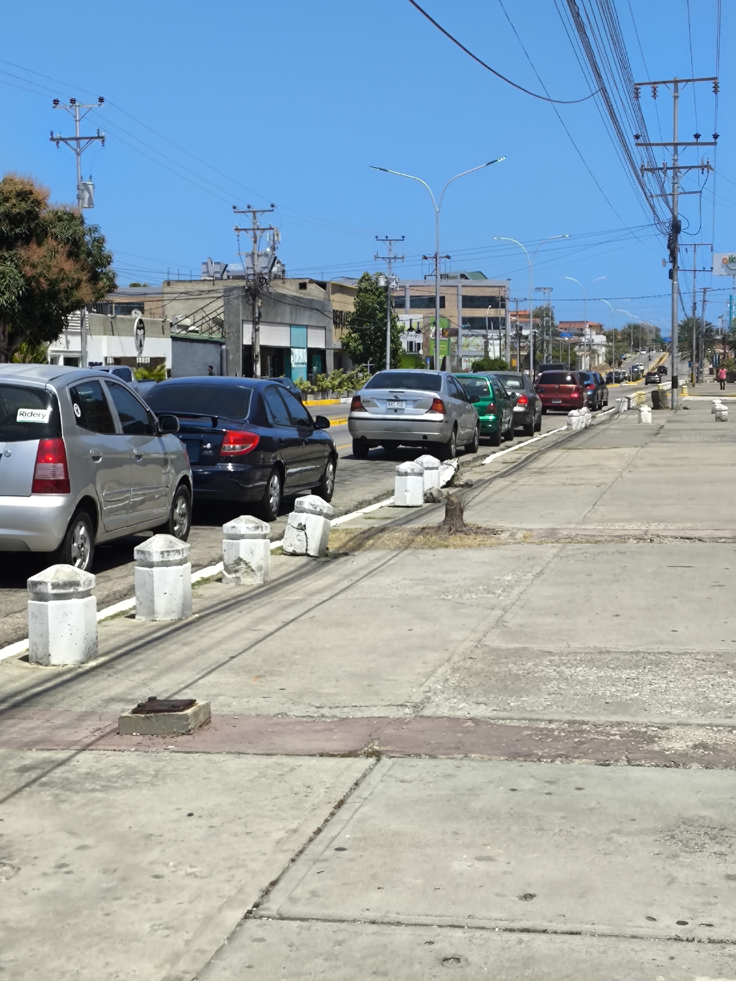 The image depicts a street in Pampatar, Venezuela, under a clear, bright blue sky. A line of parked cars occupies the right side of the road, separated from the sidewalk by a series of concrete bollards. The vehicles include a silver hatchback, a dark sedan, a silver sedan, and other various colored cars further down the street. Utility poles and power lines run along the top of the frame. The sidewalk is concrete with visible seams and variations in texture and color. A few pedestrians can be seen in the distance on the right side of the street. Buildings and vegetation are visible in the background, suggesting an urban environment. A small, square concrete structure with a metal cover is visible on the sidewalk.