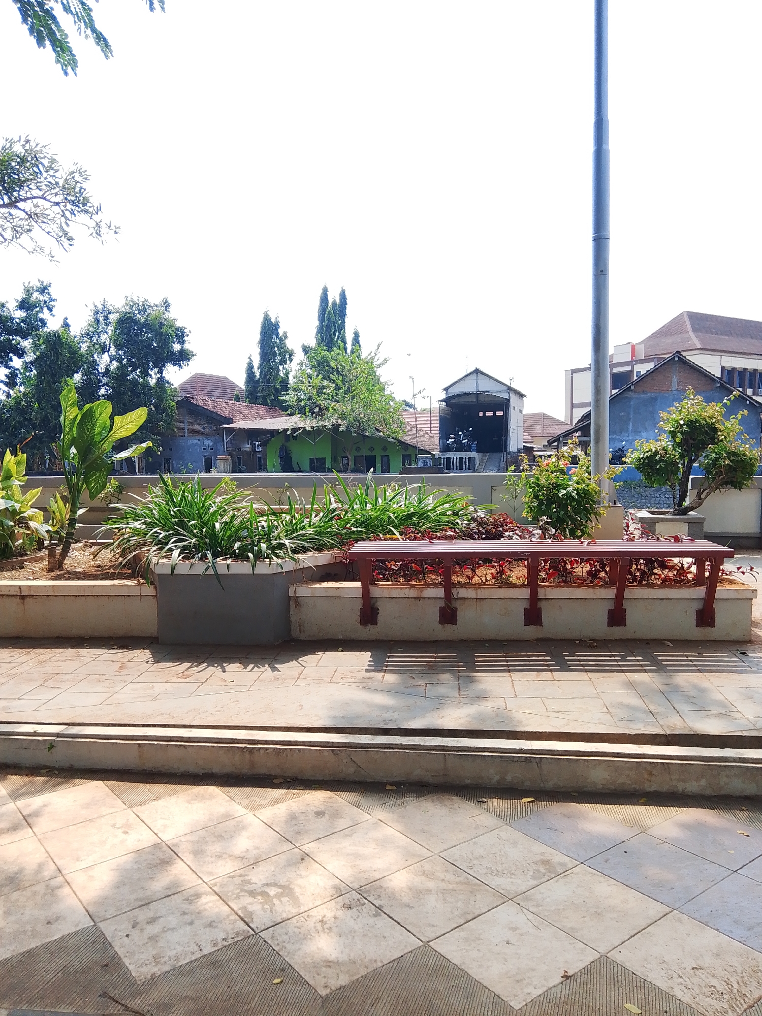 The image depicts an outdoor scene in the city of Potroyudan, Indonesia, featuring a public seating area with a planter and bench. In the foreground, a paved walkway with a diamond tile pattern is visible, leading to a raised concrete planter box. The planter contains a variety of green plants and a red wooden bench with slatted seating and metal supports. Behind the planter, a light pole stands tall against a bright sky. The background shows several buildings with different roof styles and colors, including a blue building and a green building. Trees and foliage are scattered throughout the scene, suggesting a park-like or community space. The sunlight casts shadows on the ground, indicating it is daytime. No people are present in the image.