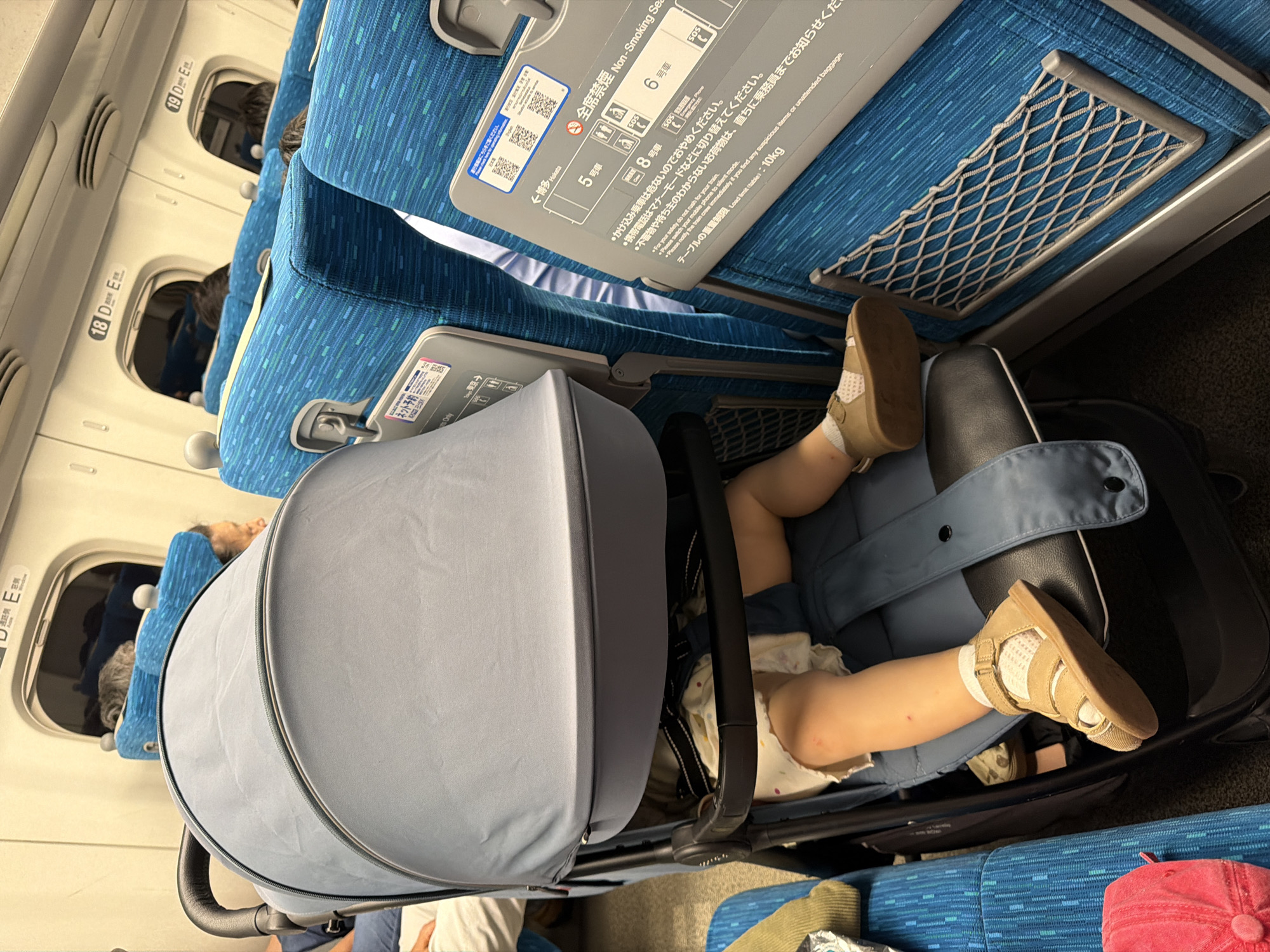 In an interior shot of a train carriage, a baby's legs are visible in a stroller. The baby wears sandals and socks. The stroller, draped with a gray canopy, is positioned next to a row of blue seats. Signs with Japanese text and safety symbols are affixed to the backs of the seats. The train appears to be in motion or stationary.
