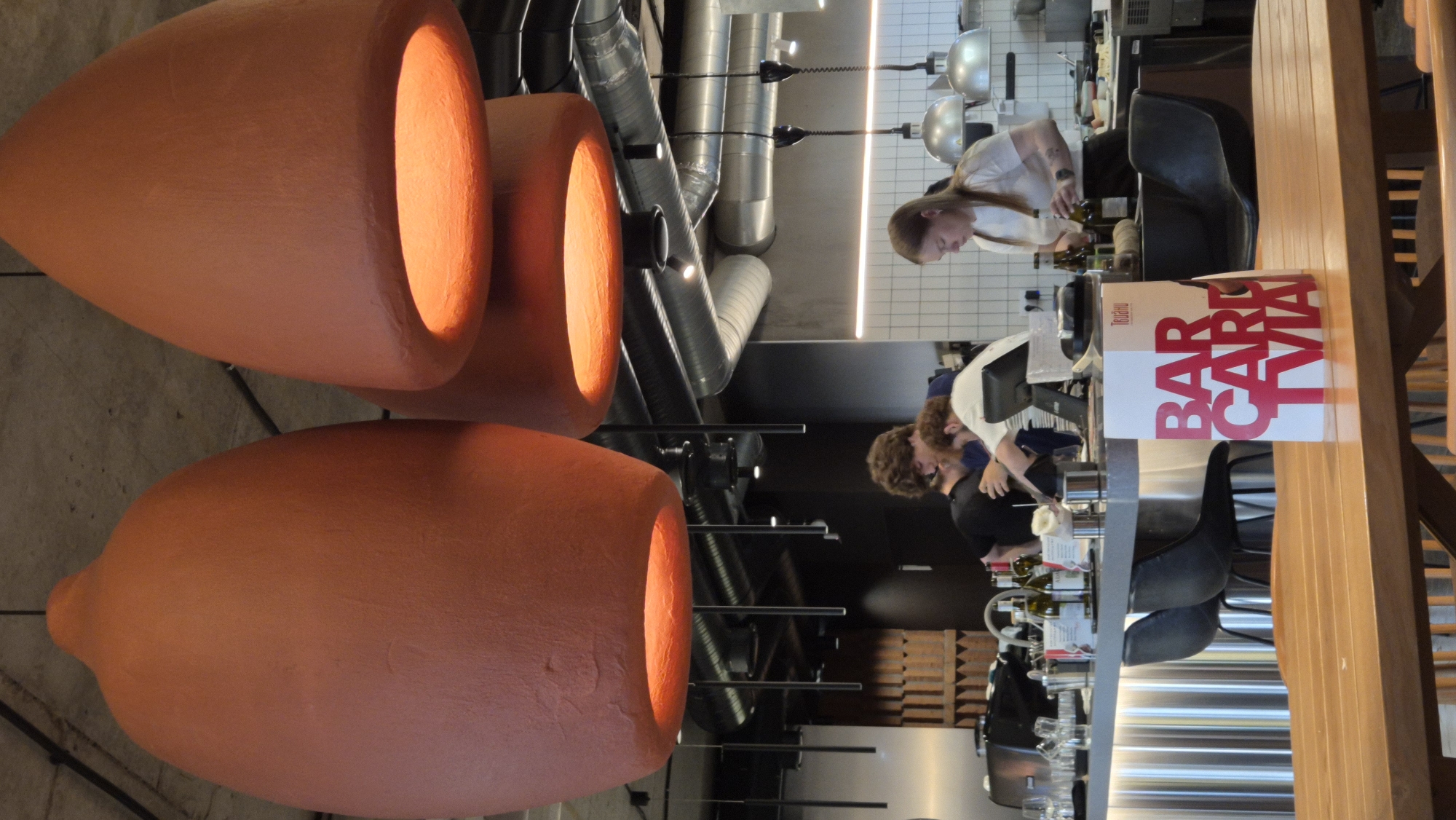 The image depicts the interior of a cafe in Kemerovo, Russia. Three large, terracotta-colored pendant lights with a textured finish hang from the ceiling, casting a warm glow downwards. In the mid-ground, a bar area is visible with a stainless steel counter. Behind the counter, three individuals are present. One person, with curly brown hair, is wearing a black t-shirt and appears to be interacting with another person who is mostly obscured and wearing a blue shirt. To the right, a young woman with blonde hair tied back, wearing a white t-shirt, is seen attending to bottles of wine. She is holding a bottle in her right hand. The background shows kitchen equipment, industrial ventilation ducts, and a tiled wall. In the foreground, a wooden table is positioned, and on it sits a white stand with red text that reads "BAR CARD TVIA." The overall environment suggests a modern and casual dining establishment.