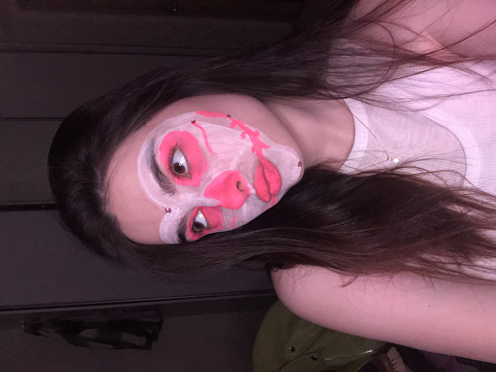 The image, captured in Noviy, Russia, presents a close-up, tilted portrait of a person with elaborate, artistic face makeup. The individual, appearing to be a young woman, has long, dark brown hair that falls across her shoulders and arm.
Her face is painted with a pale white base, over which vibrant neon pink makeup has been applied. Her eyes are dramatically outlined with bright pink, extending onto the eyelids and under the lower lash line. Her lips are also painted a glossy, vivid pink. Abstract streaks and patterns of the same neon pink run across her cheeks and forehead, creating a striking, stylized effect. Small, shimmering rhinestones or gems are strategically placed on her face, notably above her eyebrows, adding a touch of sparkle. Her brown eyes are wide open, directly engaging with the viewer, emphasizing the dramatic makeup.
She is wearing a white ribbed tank top or undershirt, and a delicate silver necklace with a small star-shaped pendant is visible around her neck. The background is dark and largely indistinct, suggesting an indoor setting with dim lighting, which makes the luminous makeup stand out. A dark, possibly wooden, surface is visible in the lower left corner, and a portion of a dark green garment or object can be seen on the right. The overall impression is one of creative expression or preparation for an event, showcasing a bold and artistic makeup design. No visible text is present in the image.