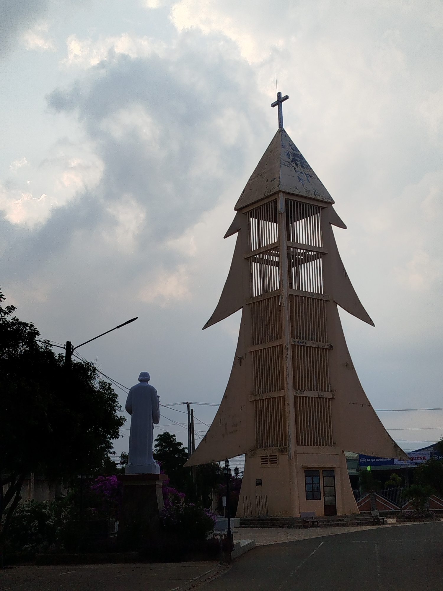 The photograph captures a scene in Suối Thông C2, Vietnam, featuring a distinctive bell tower of a local church. The tower, painted in a light tan color, is designed with a unique shape reminiscent of a stylized Christmas tree, complete with layered triangular elements. At its apex sits a Christian cross. The tower's structure is partially enclosed with vertical bars.
Adjacent to the tower is a statue of a figure in flowing robes, potentially a religious figure, standing on a raised plinth surrounded by vegetation and flowering plants. The statue is facing away from the camera. The area around the tower and statue appears to be a paved plaza or open space.
The sky is overcast with various layers of clouds, creating an atmospheric backdrop. Utility poles and power lines are visible in the background. Distant buildings and signage with text are also noticeable, though details are less clear. Overall, the scene is quiet and still, emphasizing the religious and commemorative nature of the setting.